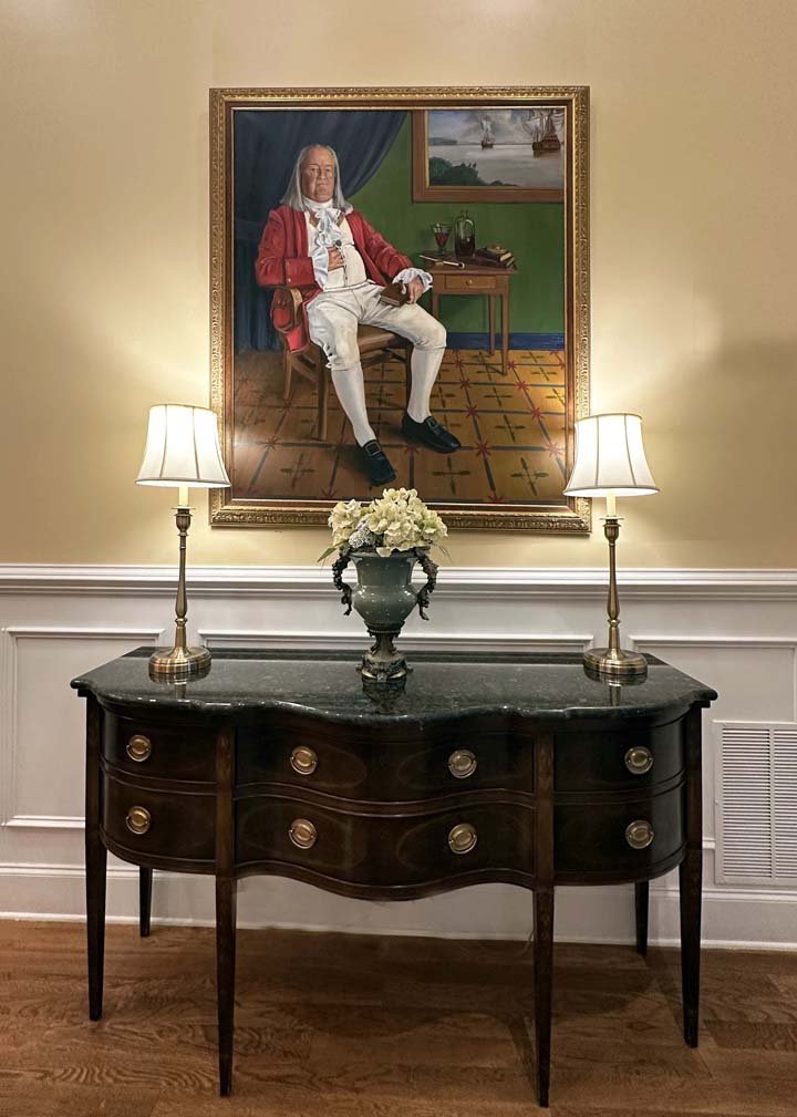A portrait of a man in a red coat sits above a wooden desk with a vase of flowers and two lamps.