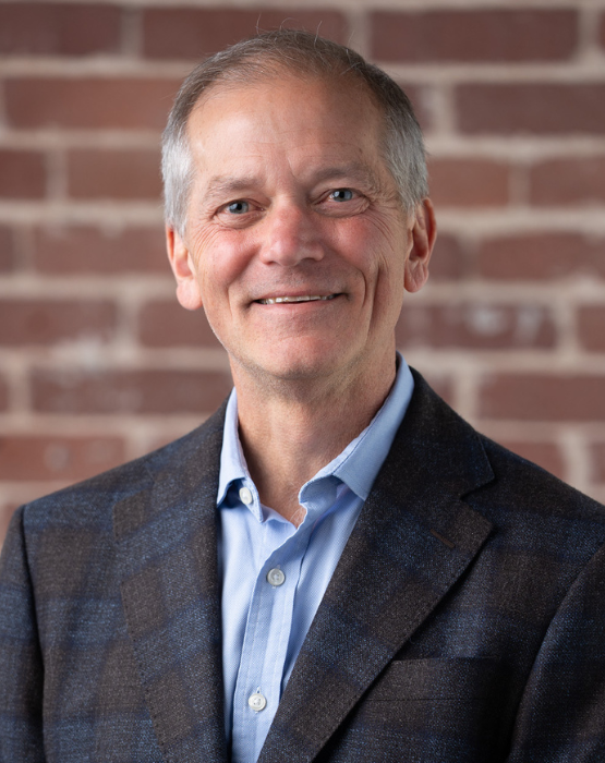 A smiling man in a blue shirt and gray blazer stands in front of a brick wall