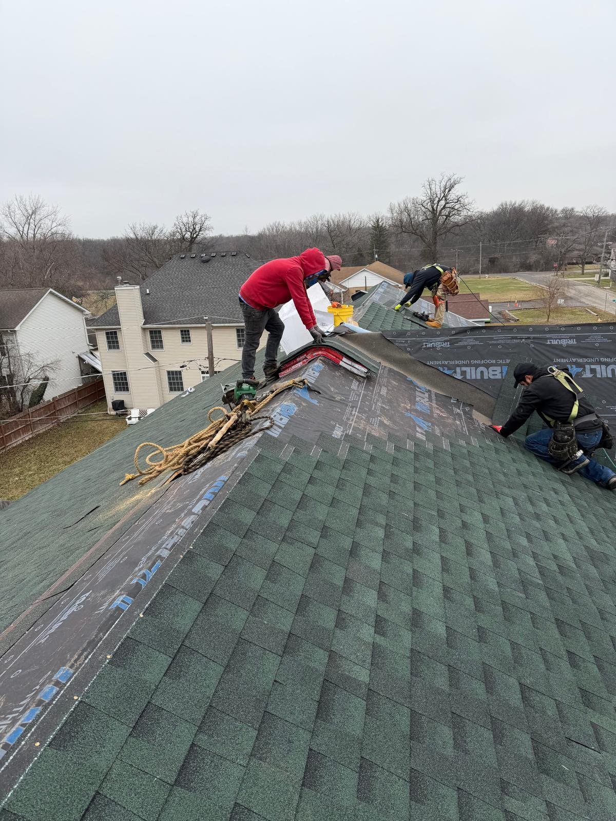 Three workers on a roof installing shingles with a house and trees in the background