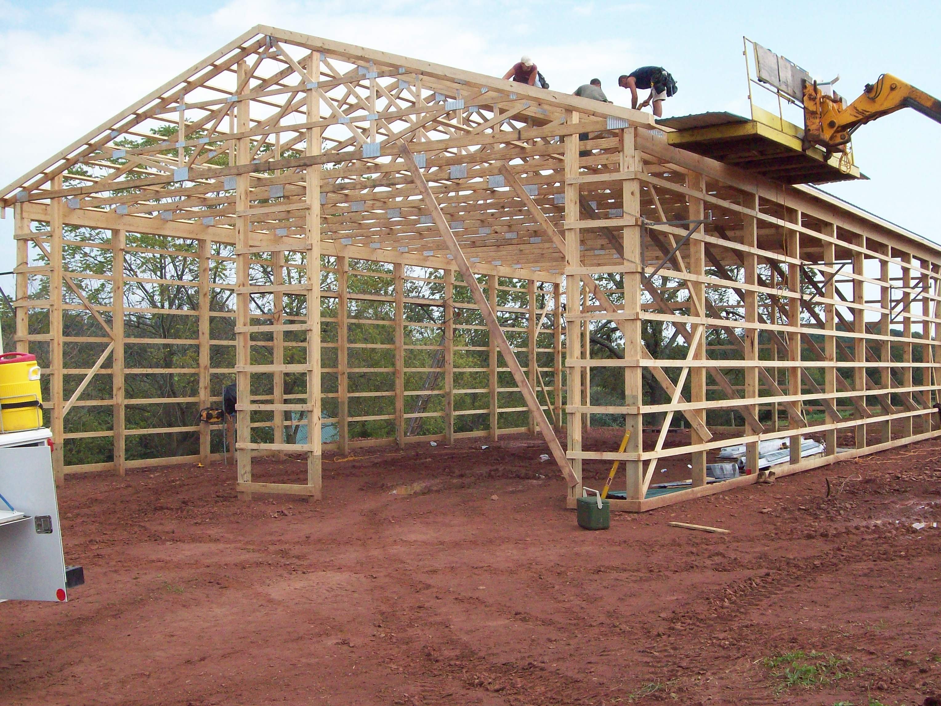Three workers constructing a wooden frame for a barn on a dirt field under a cloudy sky.