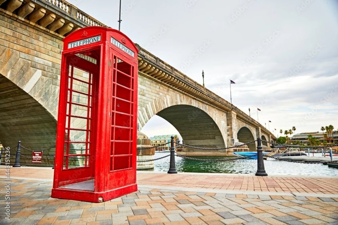 A red British telephone booth stands on a brick walkway near a bridge over a body of water.