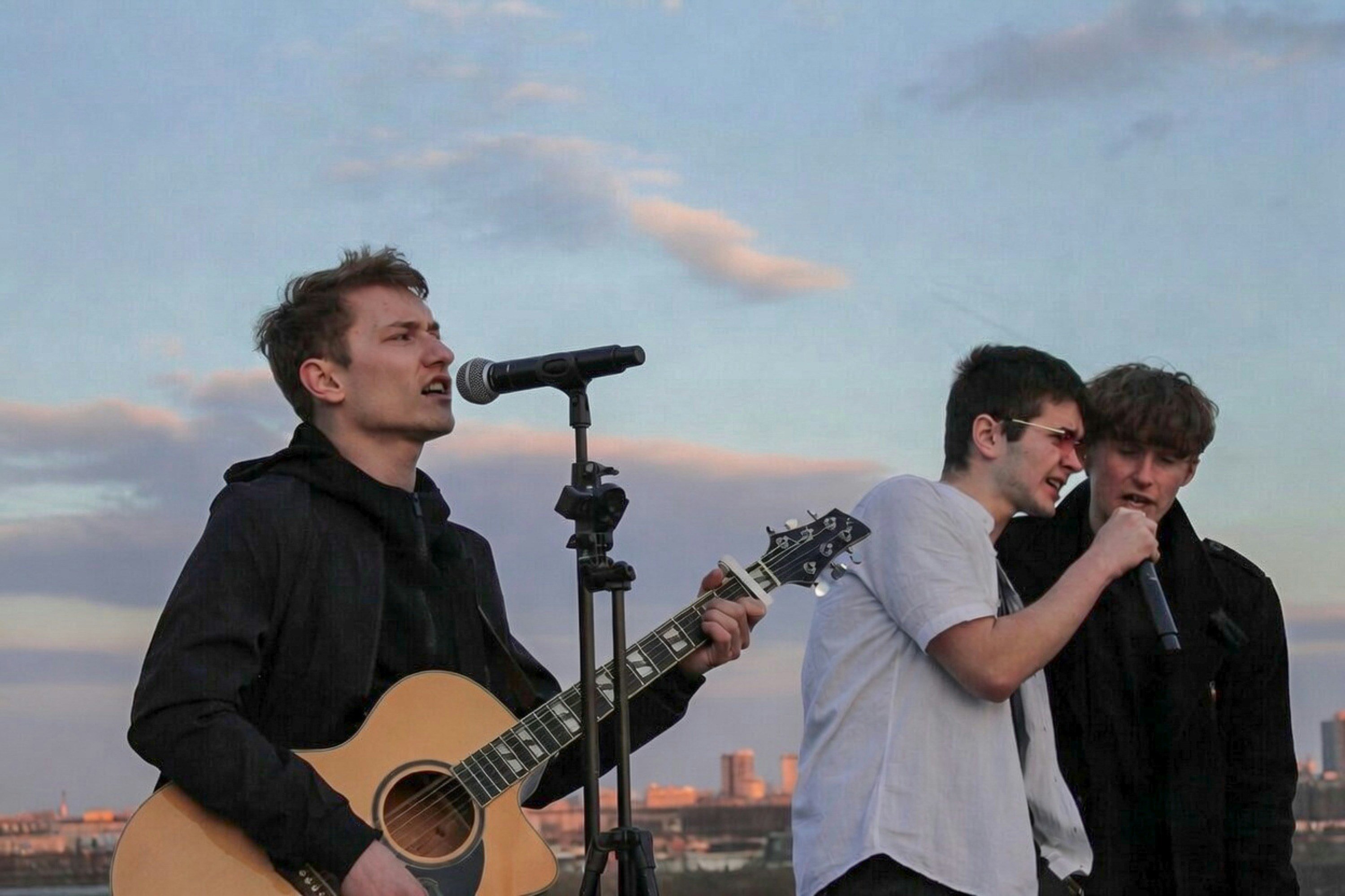 Three young men performing on stage with one playing guitar and singing, another singing into a microphone, and the third sin
