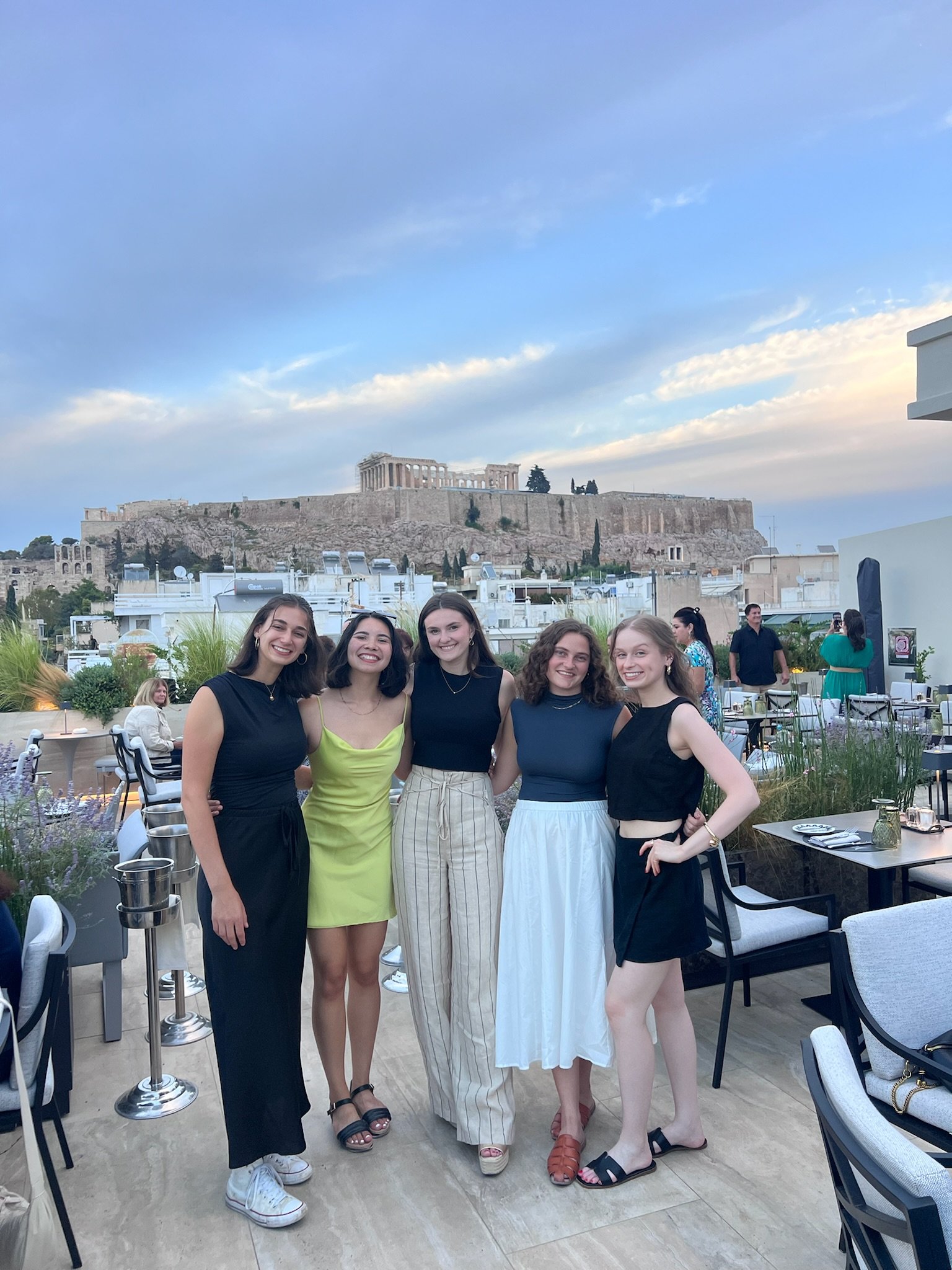 Five women in evening wear pose for a photo on a rooftop terrace overlooking the Acropolis in Athens.
