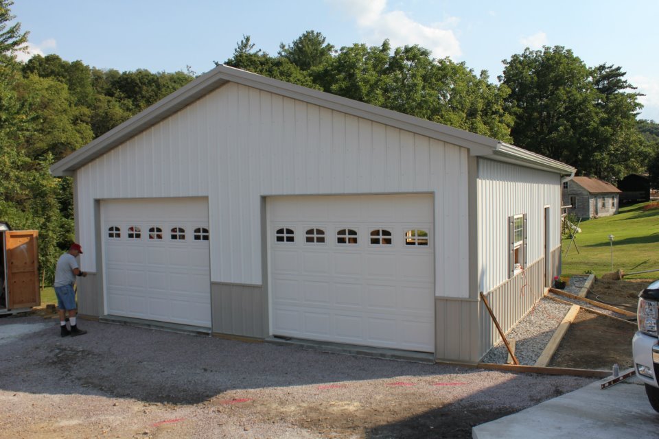 A man stands in front of a white garage with two doors and a gray roof, surrounded by trees and a grassy field.