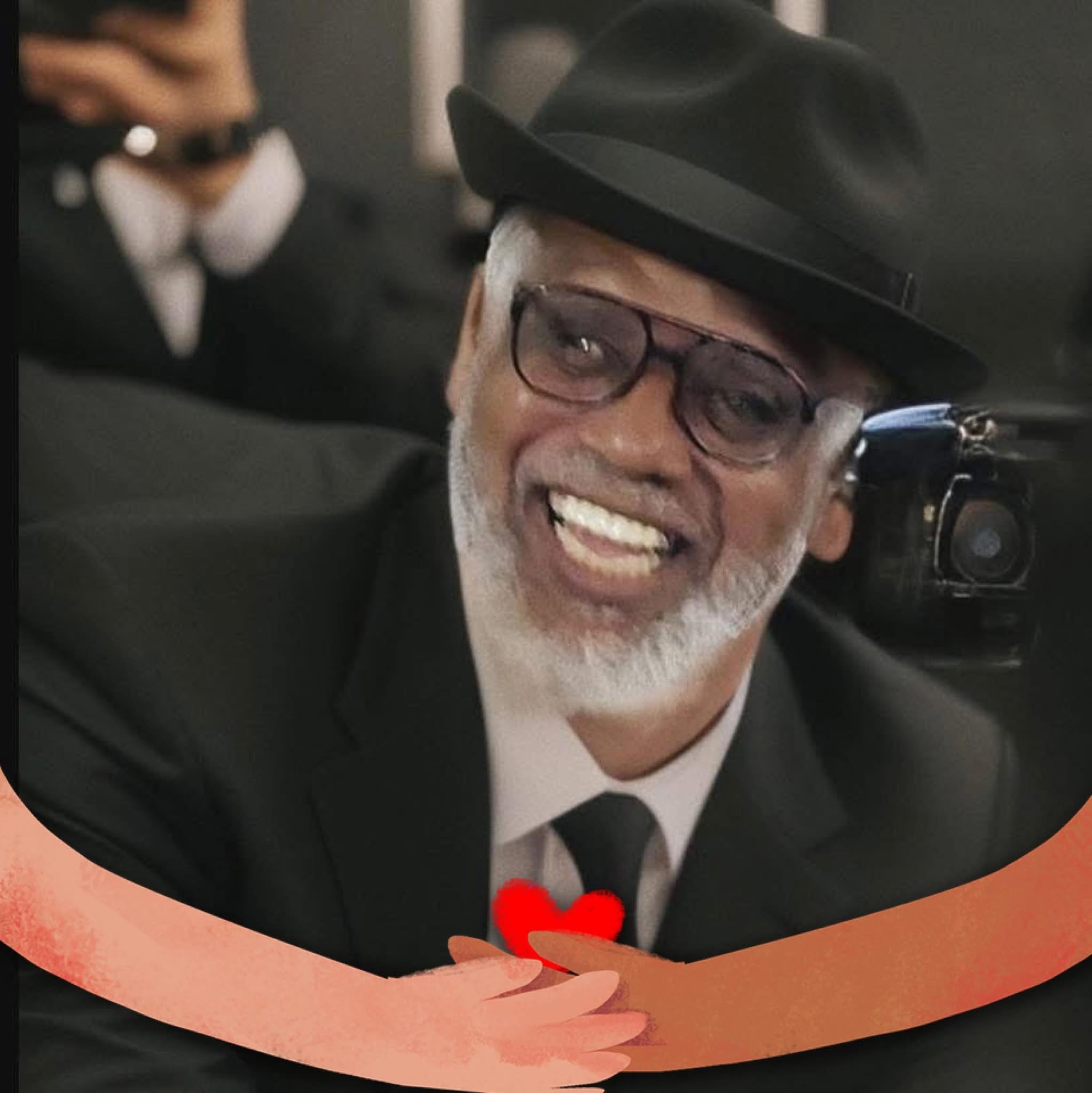 An older man in a black suit and hat smiles warmly, with a red heart drawn around his hands