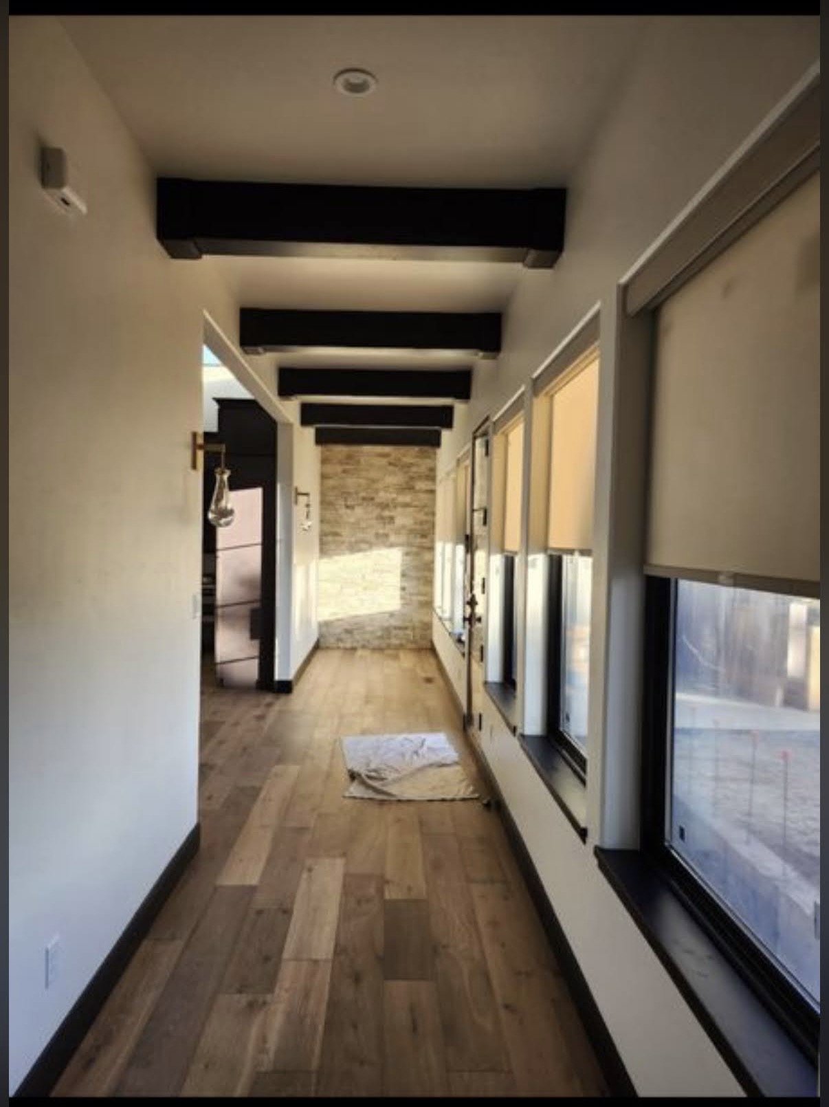 A hallway with wooden floors and white walls, featuring a brick wall and large windows with blinds.