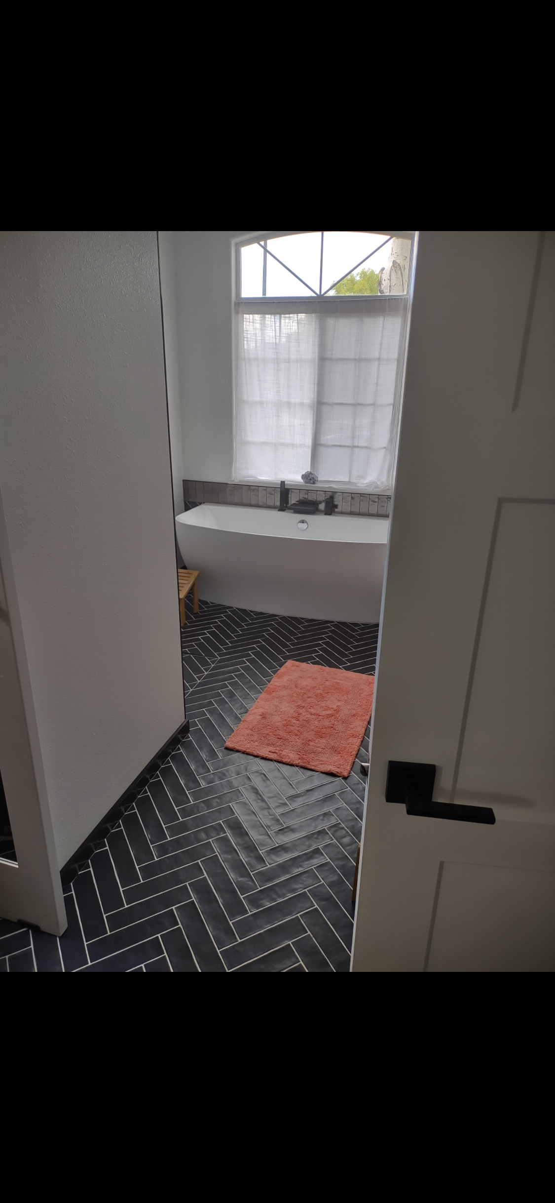 A modern bathroom with a white bathtub, black tiled floor, and a window with white curtains.