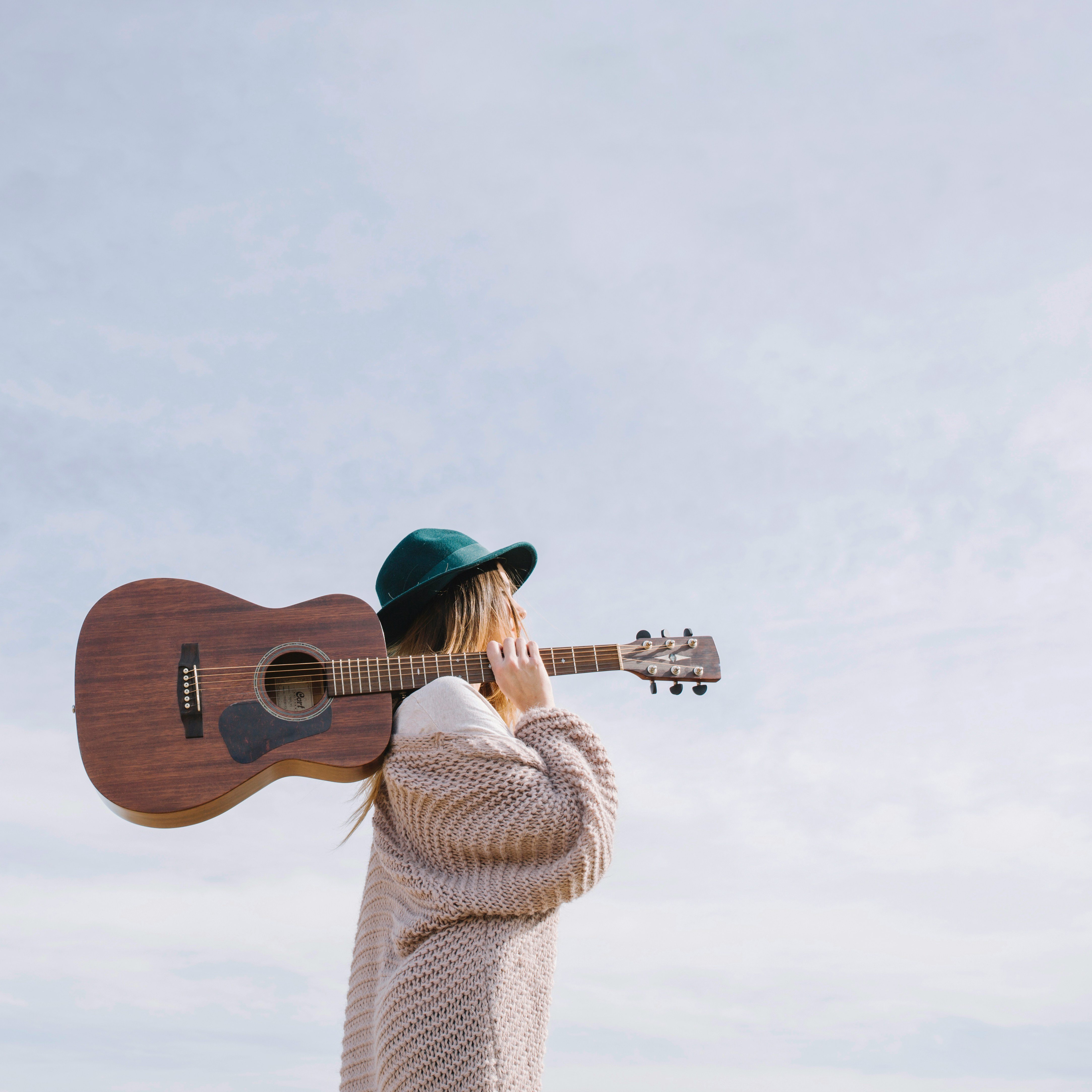 A woman wearing a green hat and a beige sweater holds a guitar over her shoulder.