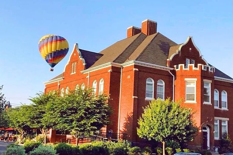 A colorful hot air balloon floats above a red brick building with arched windows and a brown roof.