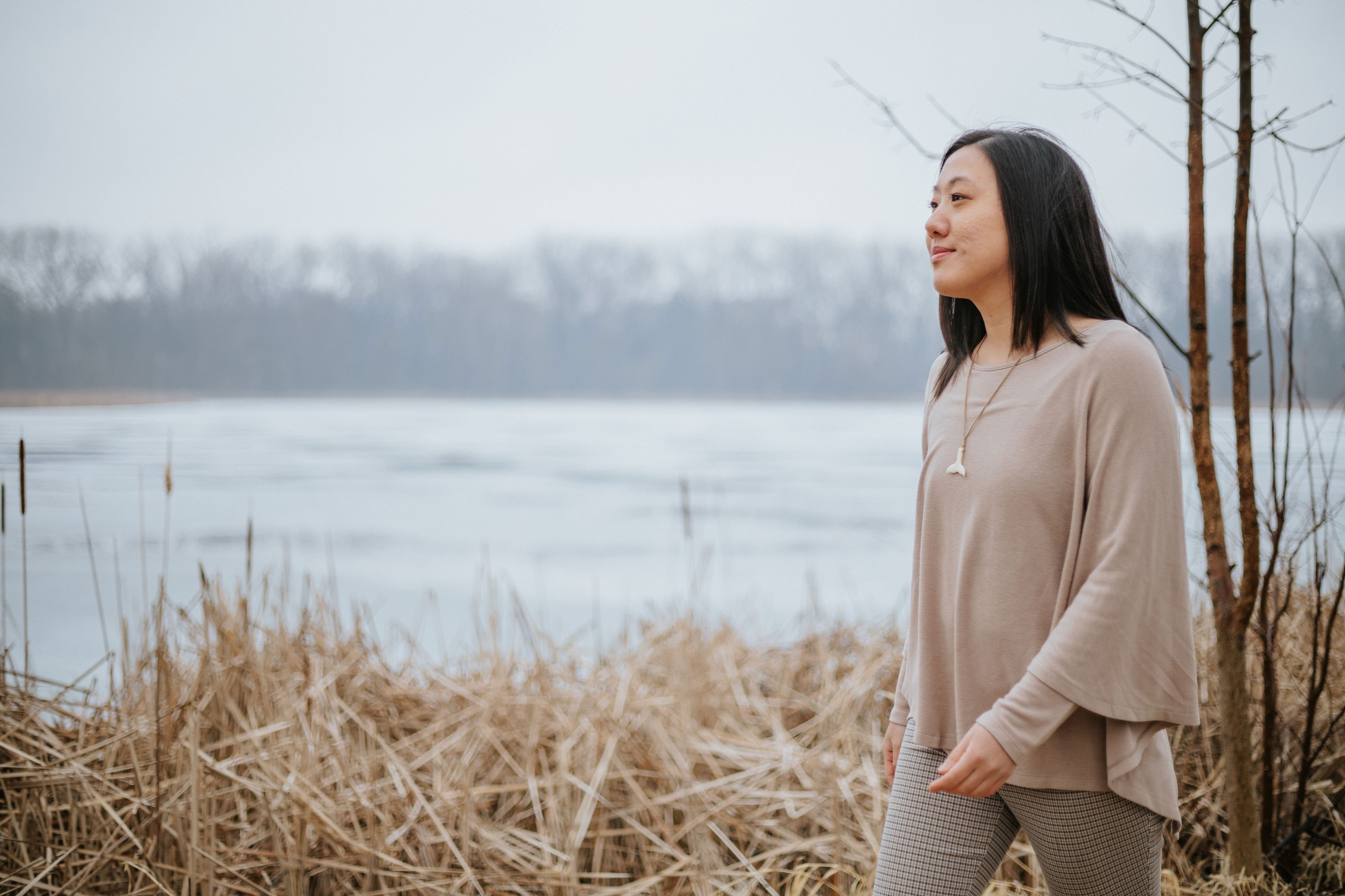 A woman stands by a lake in winter, wearing a beige top and plaid pants, looking out at the water.