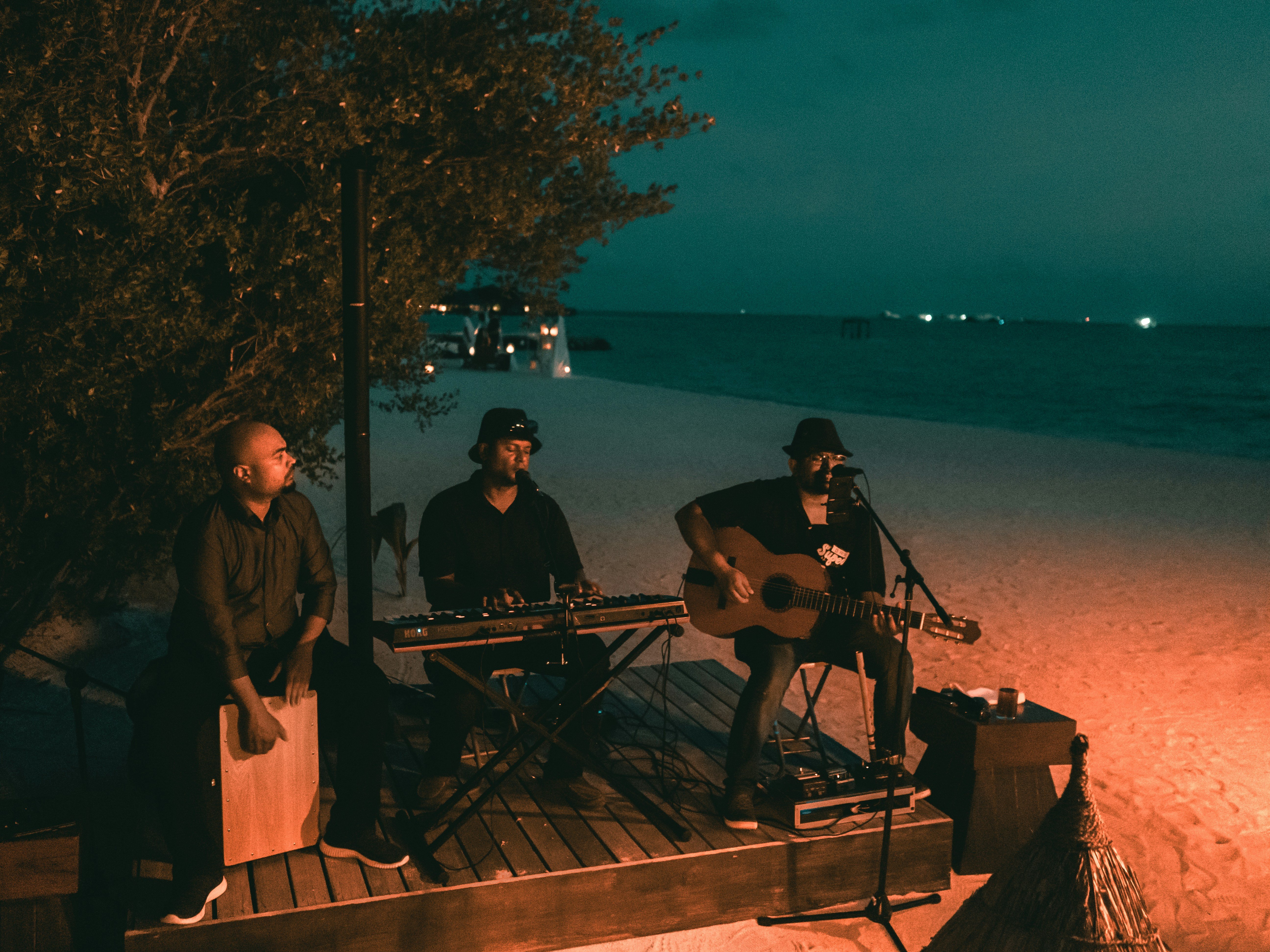 Three musicians perform on a beach at night, with one playing guitar and singing, another playing keyboard, and the third pla