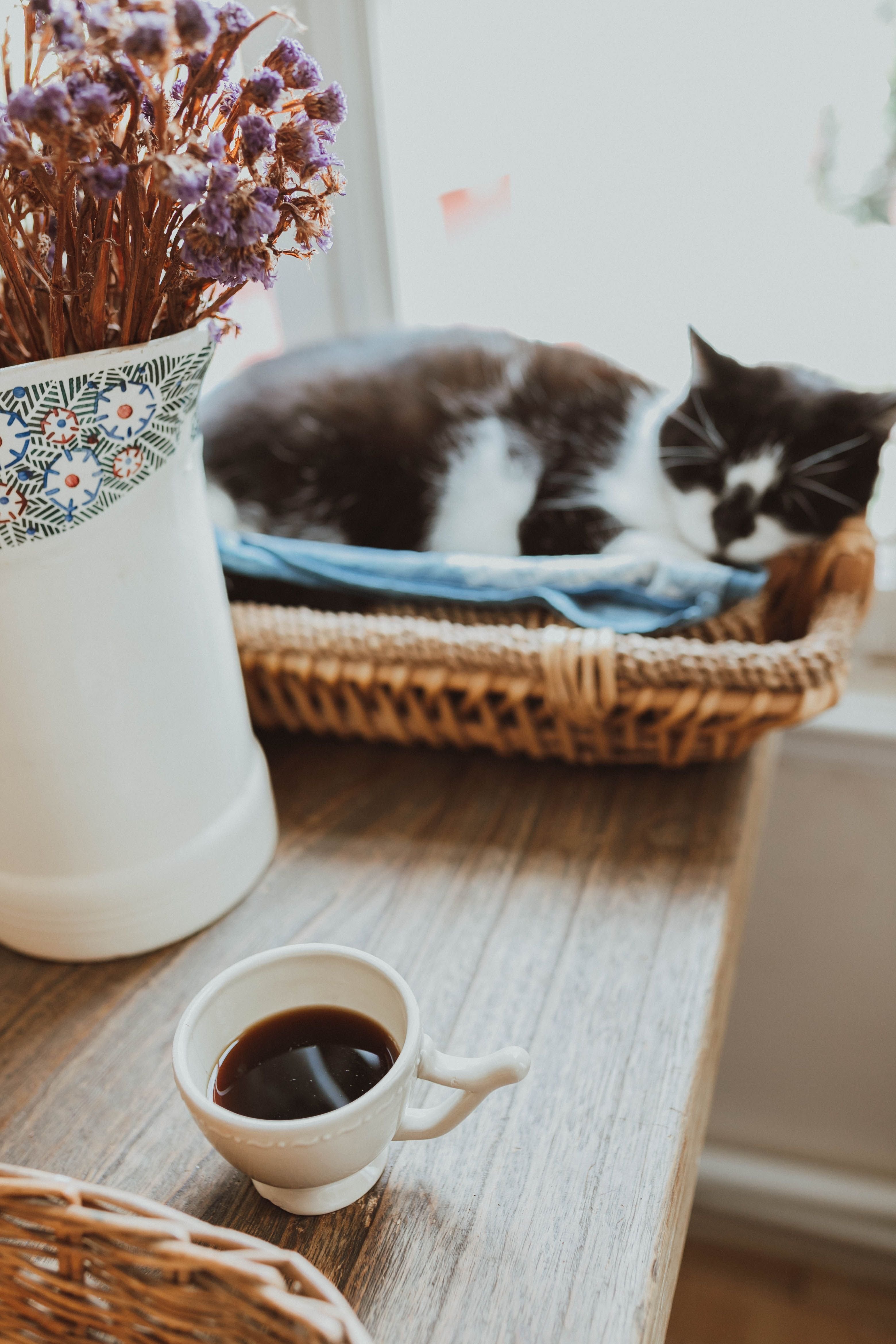A black and white cat sleeping in a basket next to a cup of coffee and a vase of dried flowers.