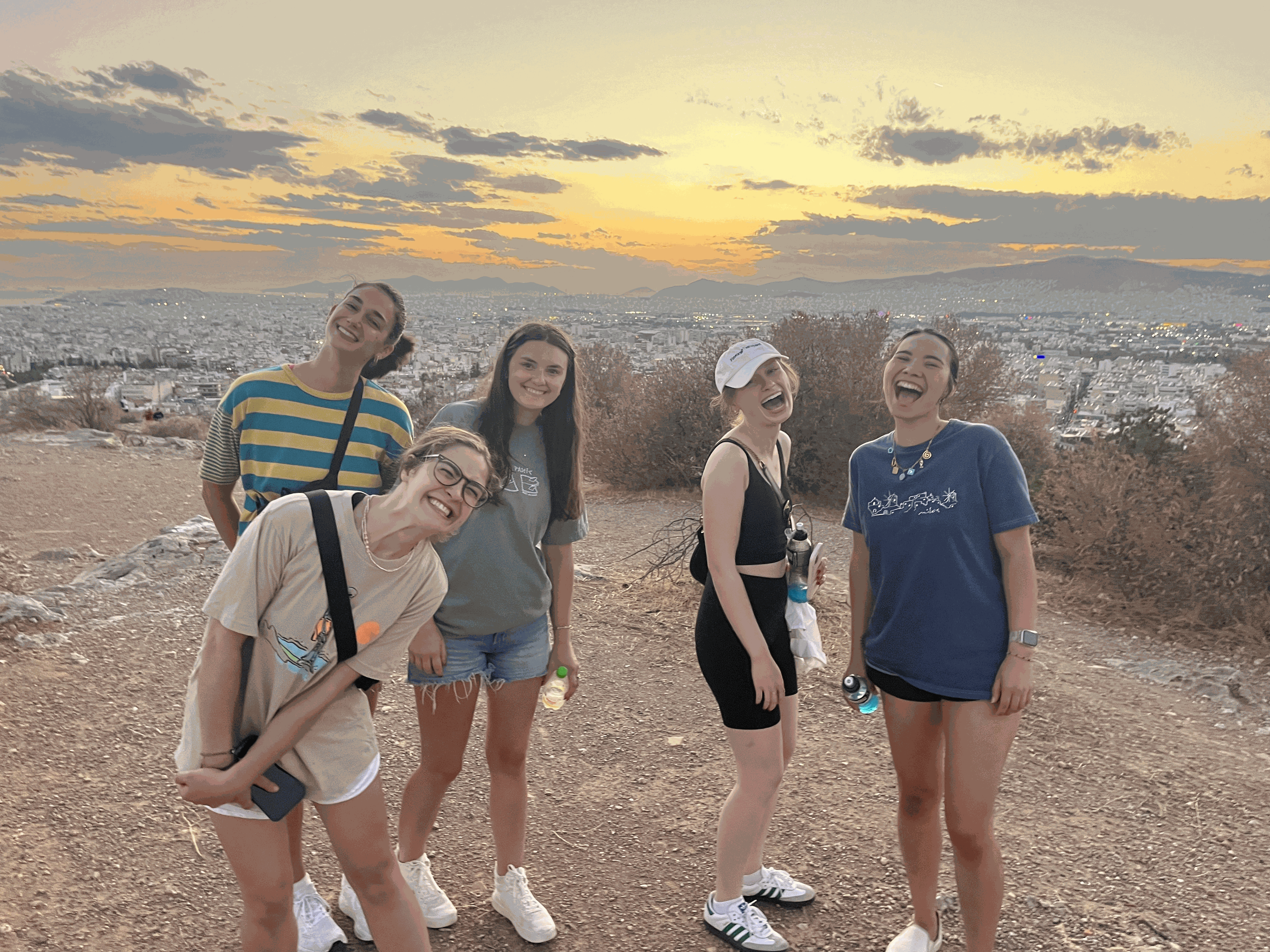 Five women standing on a hill overlooking a city at sunset, smiling and posing for a photo