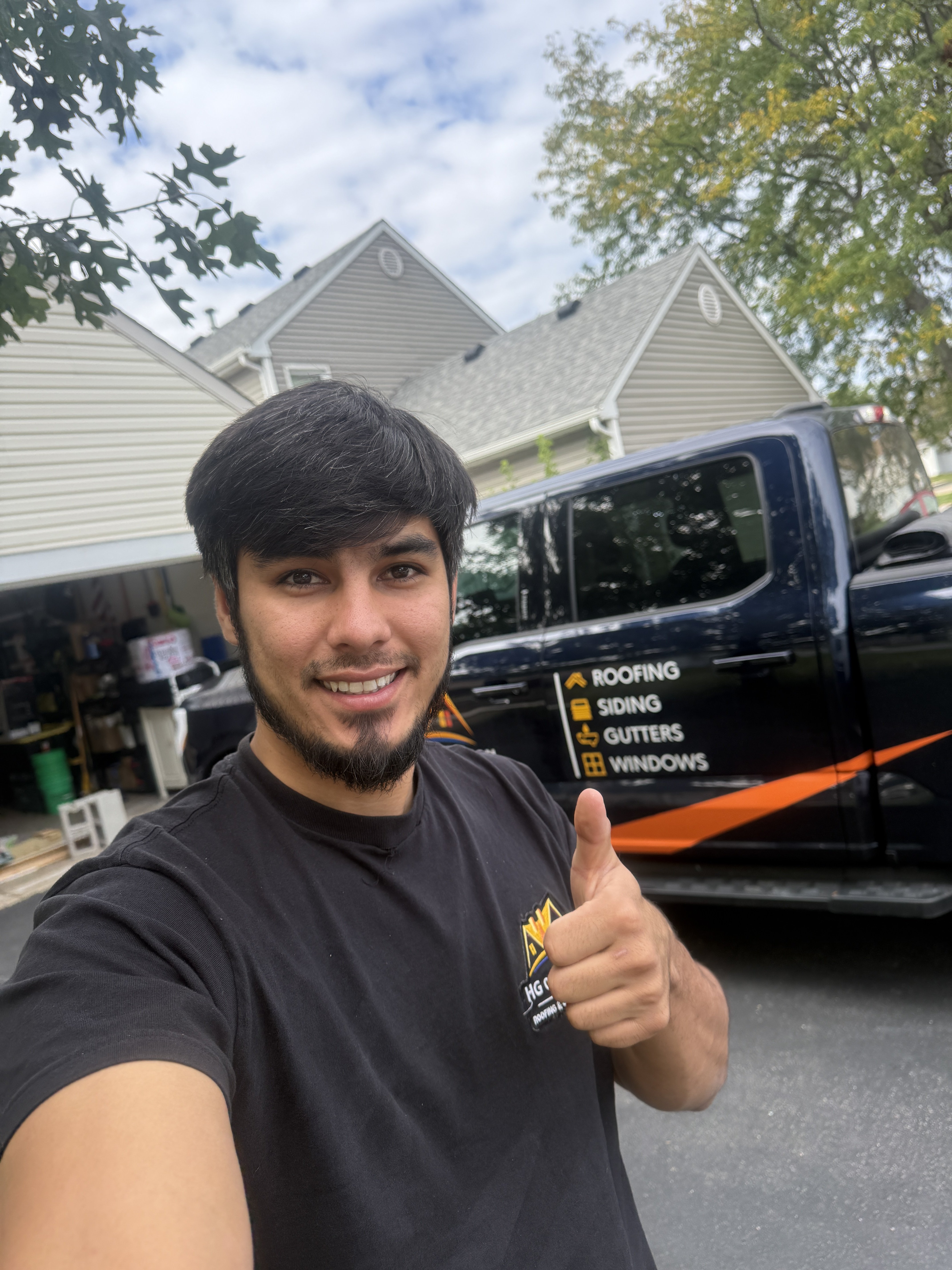 A smiling man in a black t-shirt giving a thumbs up in front of a house and a truck with roofing services branding.
