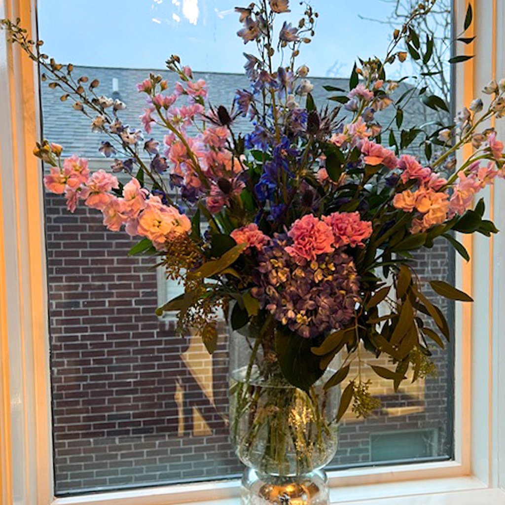 A vibrant bouquet of pink, purple, and blue flowers in a glass vase on a windowsill