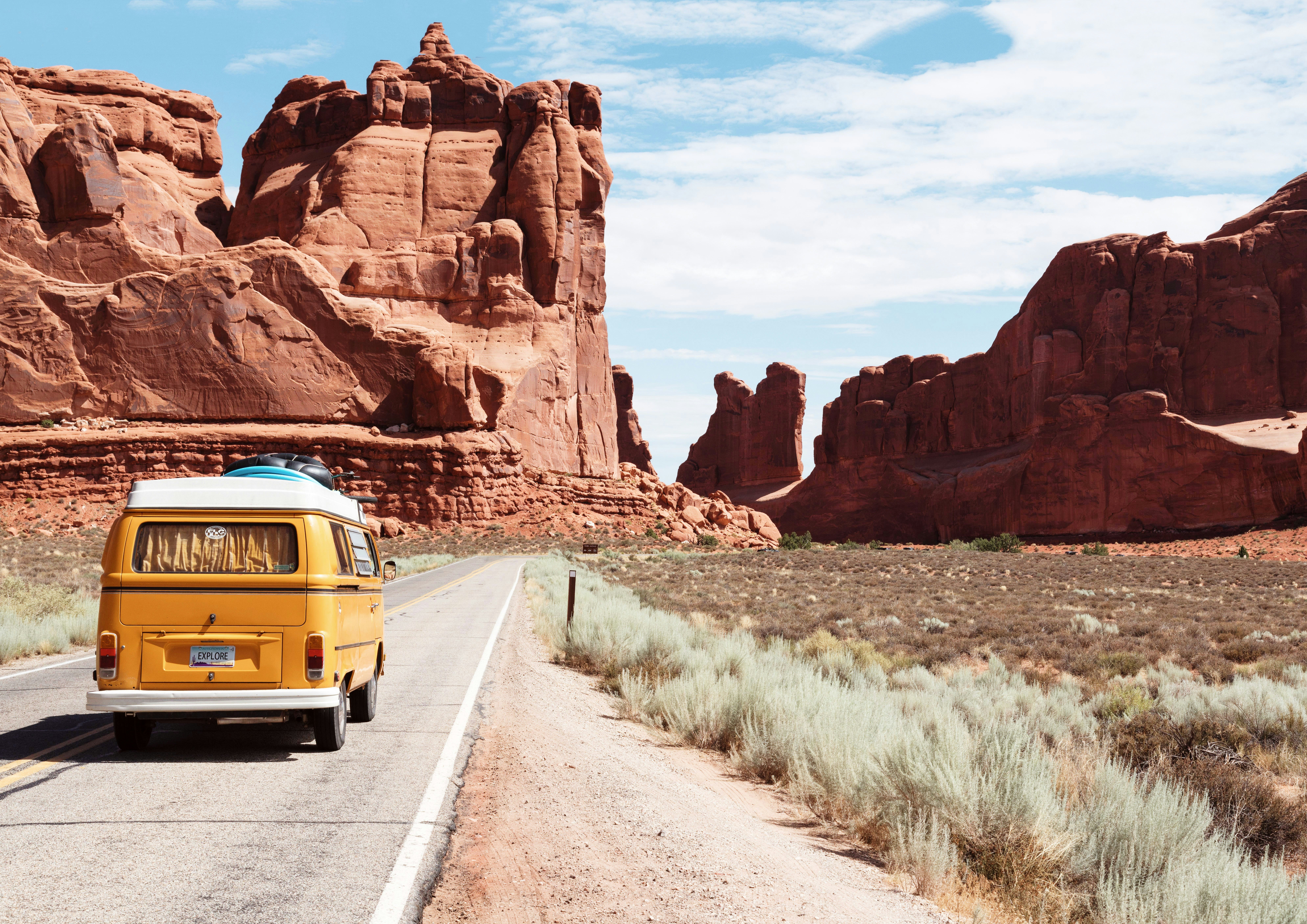 A yellow VW camper van drives down a road with red rock formations in the background.