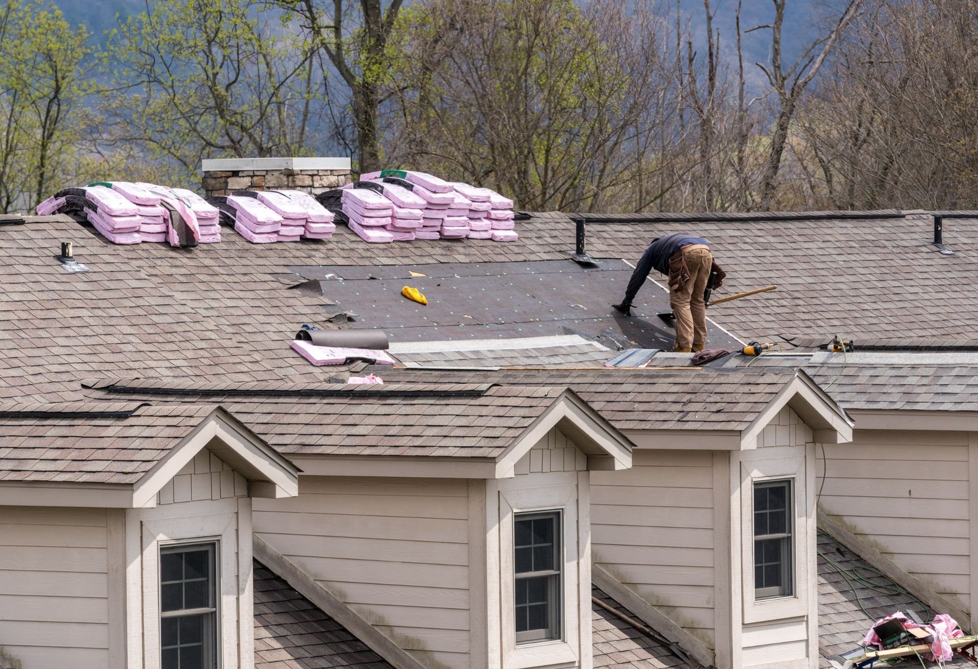 Roofer on a residential roof installing new shingles with pink insulation bundles stacked nearby