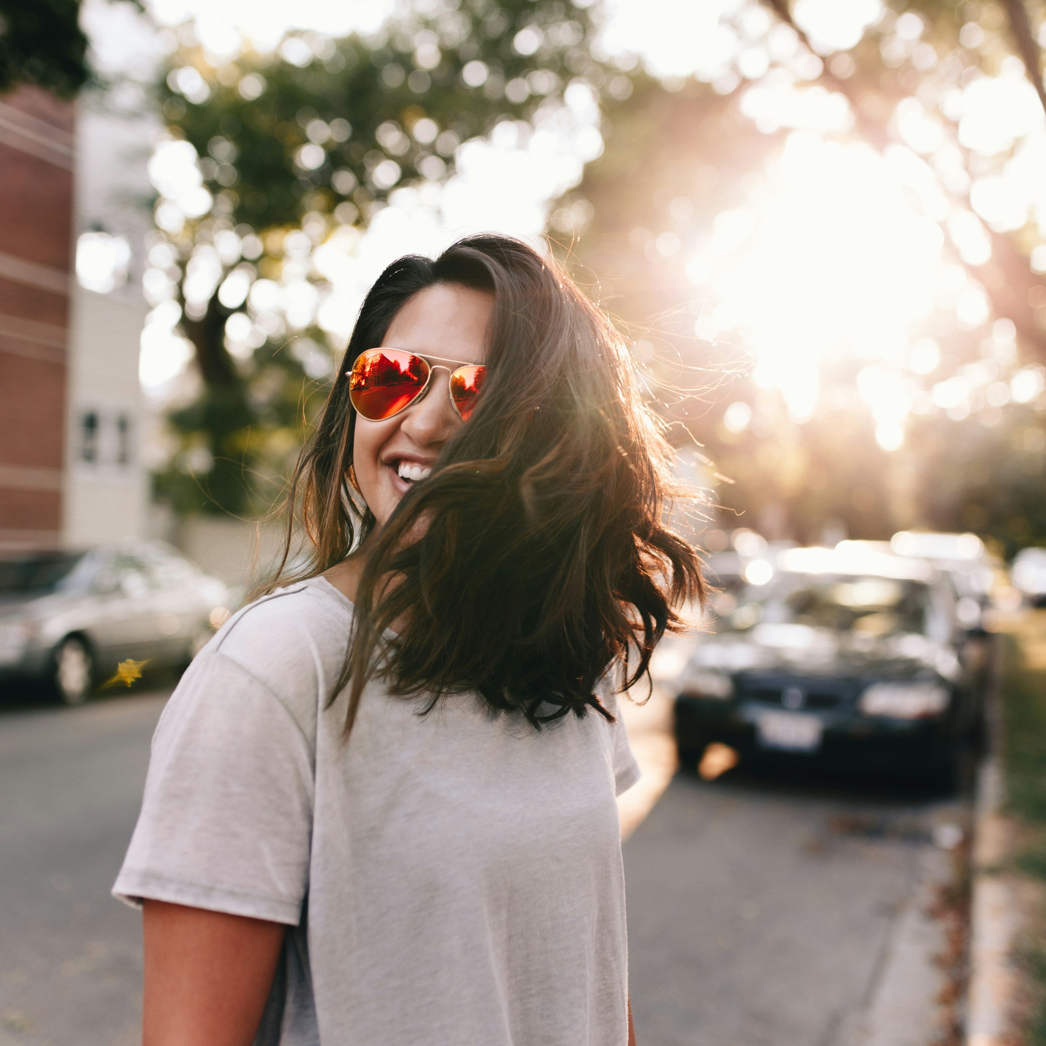 A woman with long hair wearing sunglasses and a white t-shirt smiles on a sunny street.