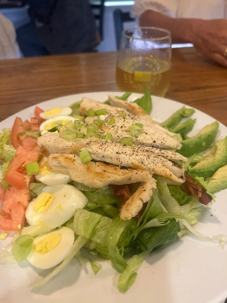 A white plate with a chicken salad with bacon, eggs, and avocado on a wooden table