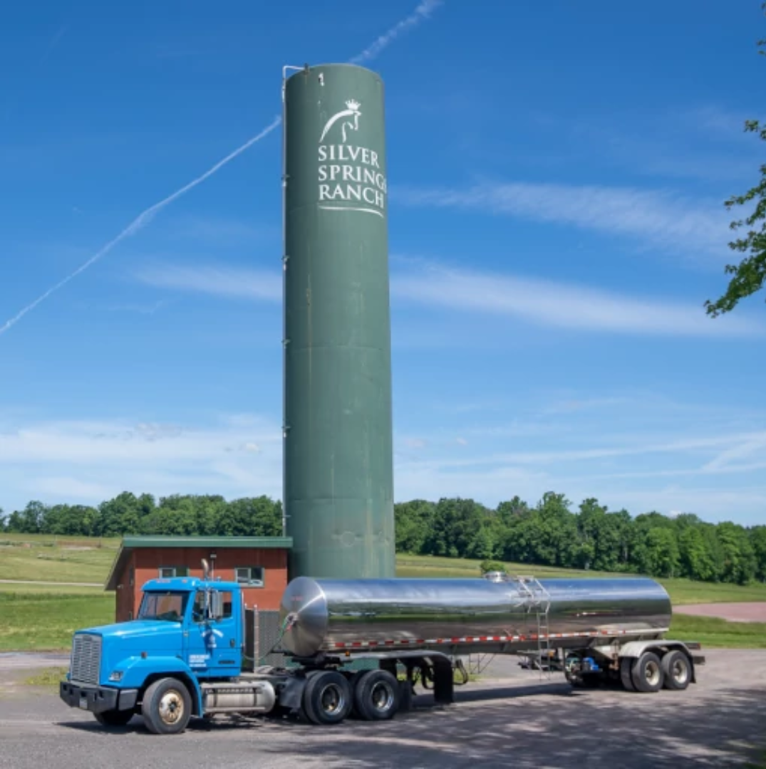 A blue truck with a large silver tank attached is parked in front of a green silo