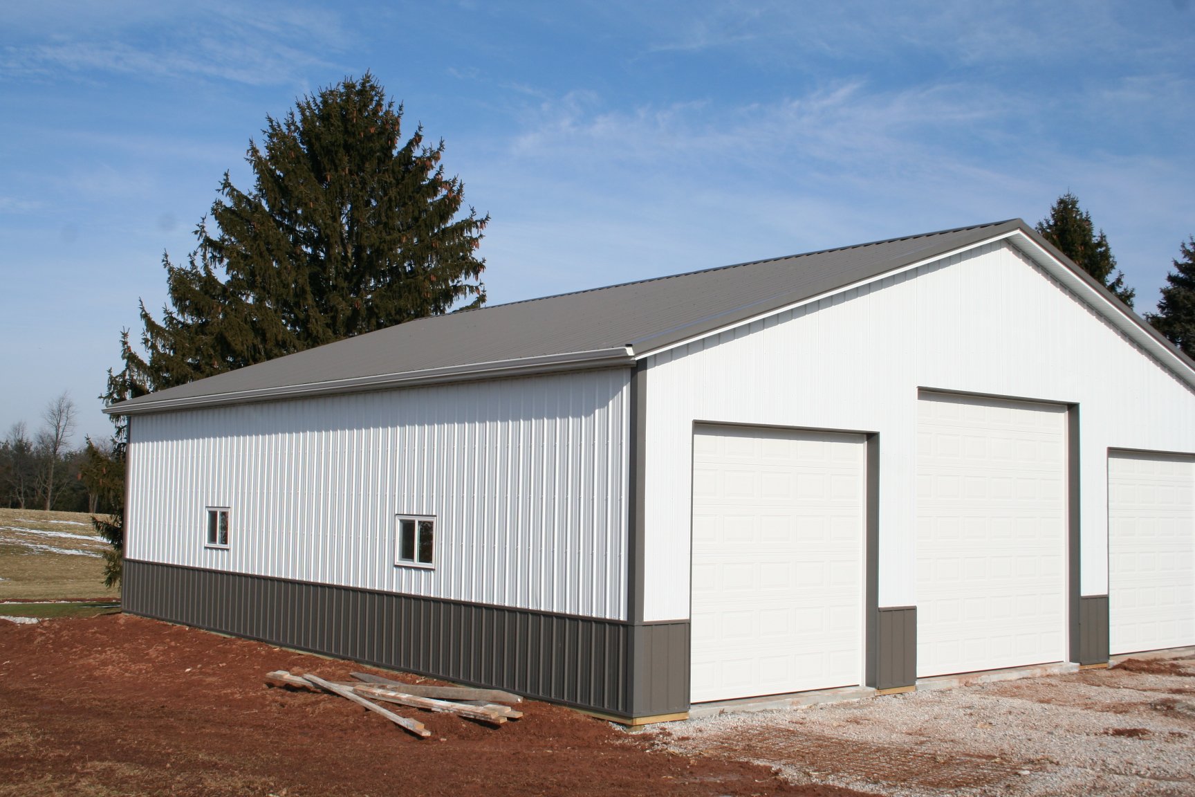 A white metal building with a gray roof and three garage doors, surrounded by trees and a field