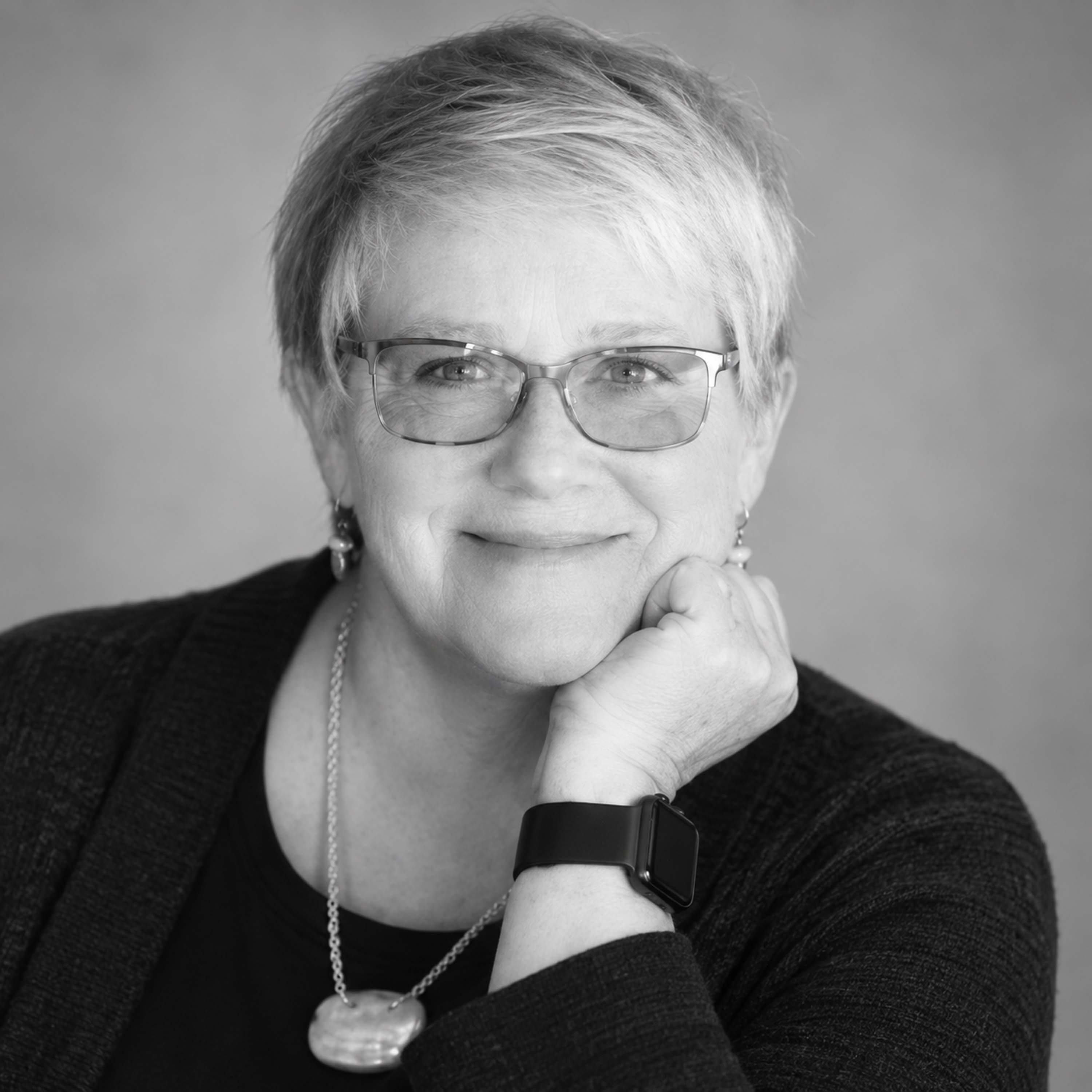 A black and white headshot of a woman with short hair, glasses, and a necklace