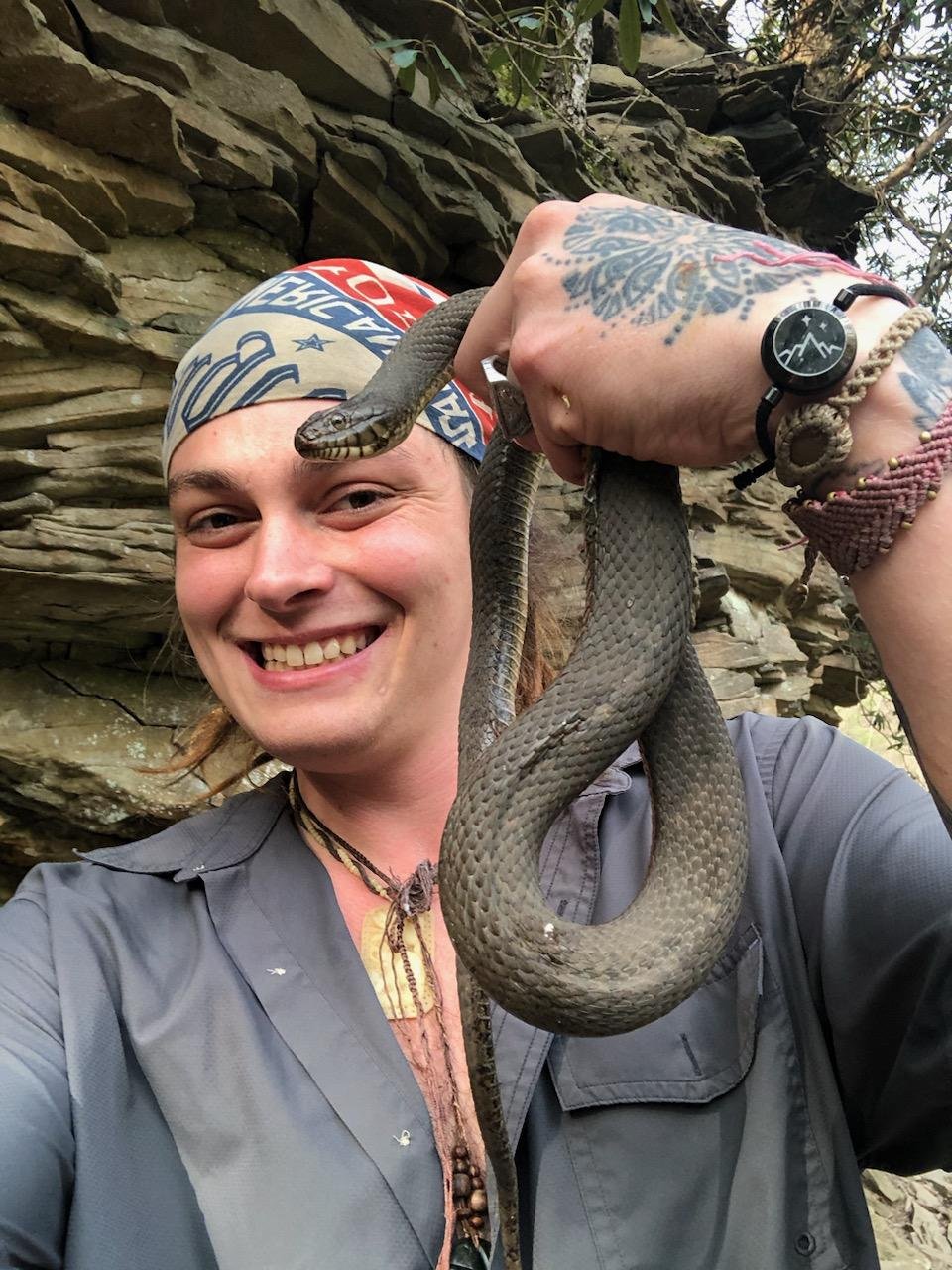 A smiling person with tattoos holds a gray snake in front of a rock wall