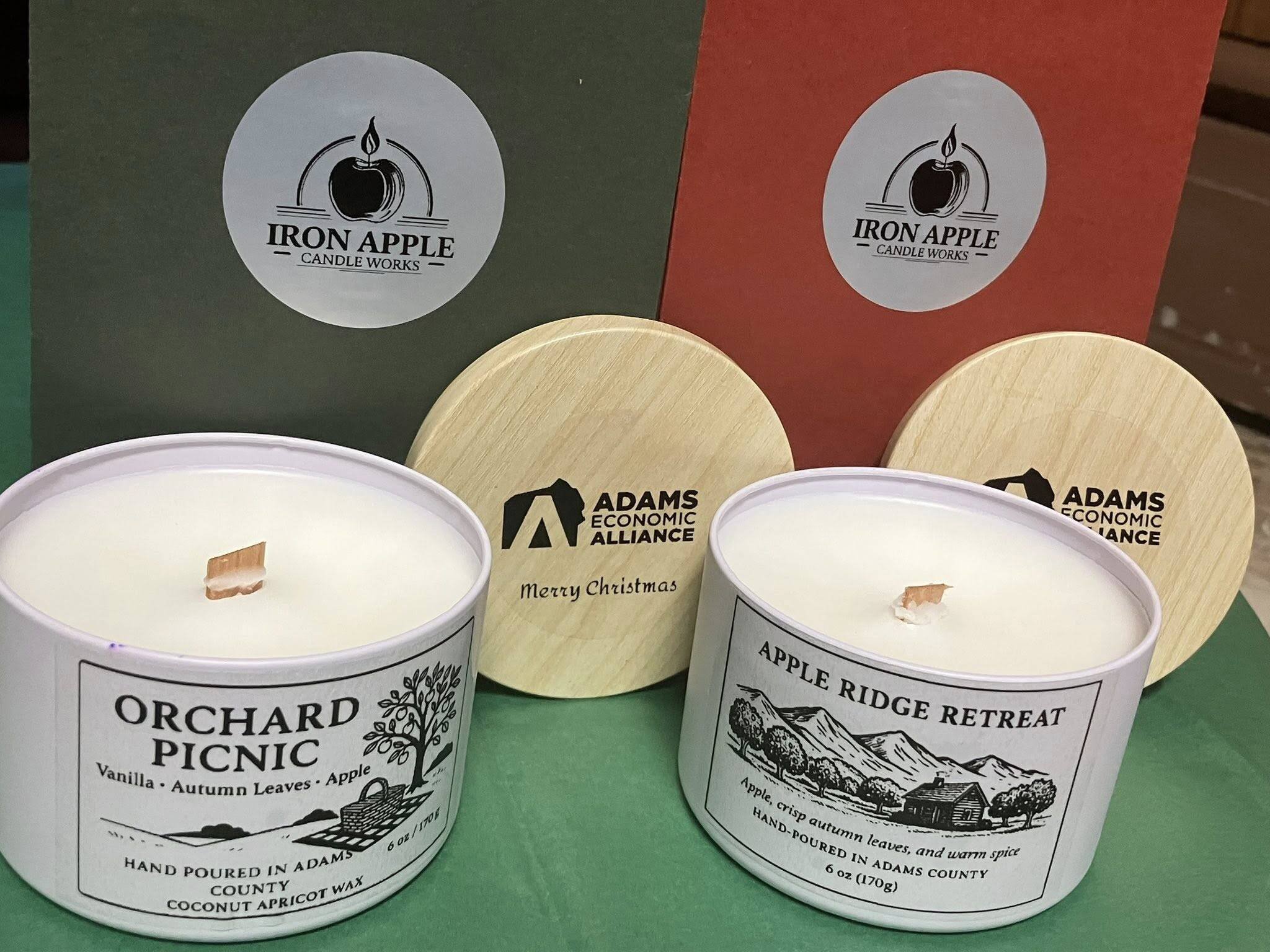 Two Iron Apple Candle Works candles on a green surface with wooden coasters and boxes