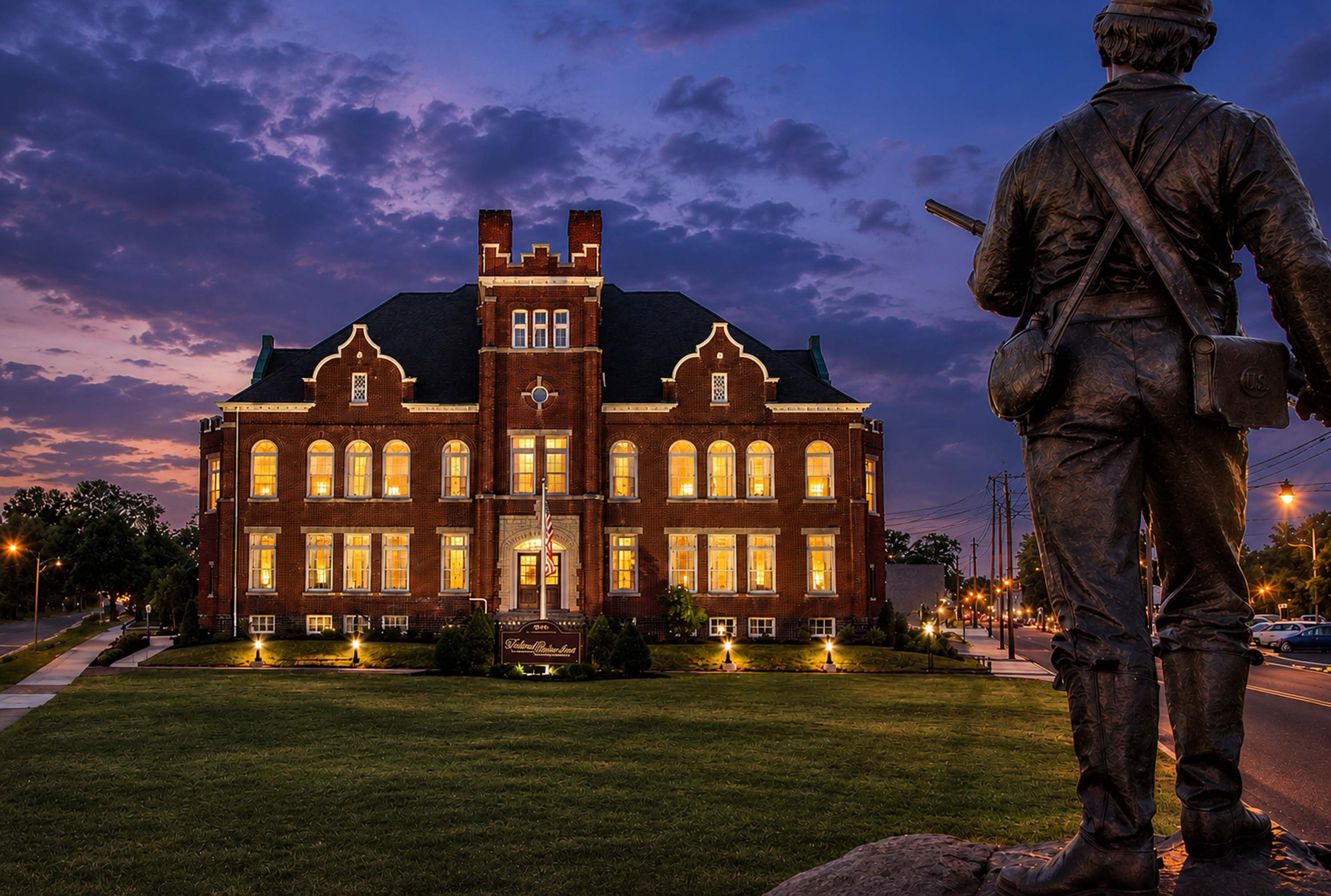 A statue of a soldier stands in front of a historic brick building at dusk.