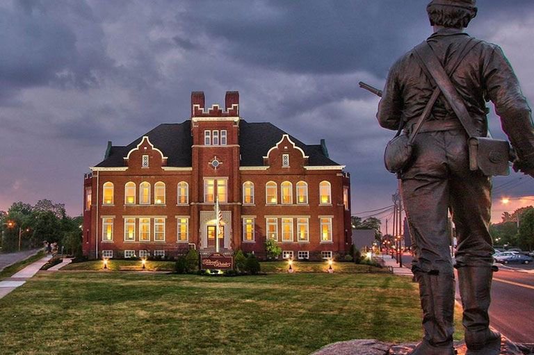 A statue of a soldier stands in front of a historic red brick building with many windows.