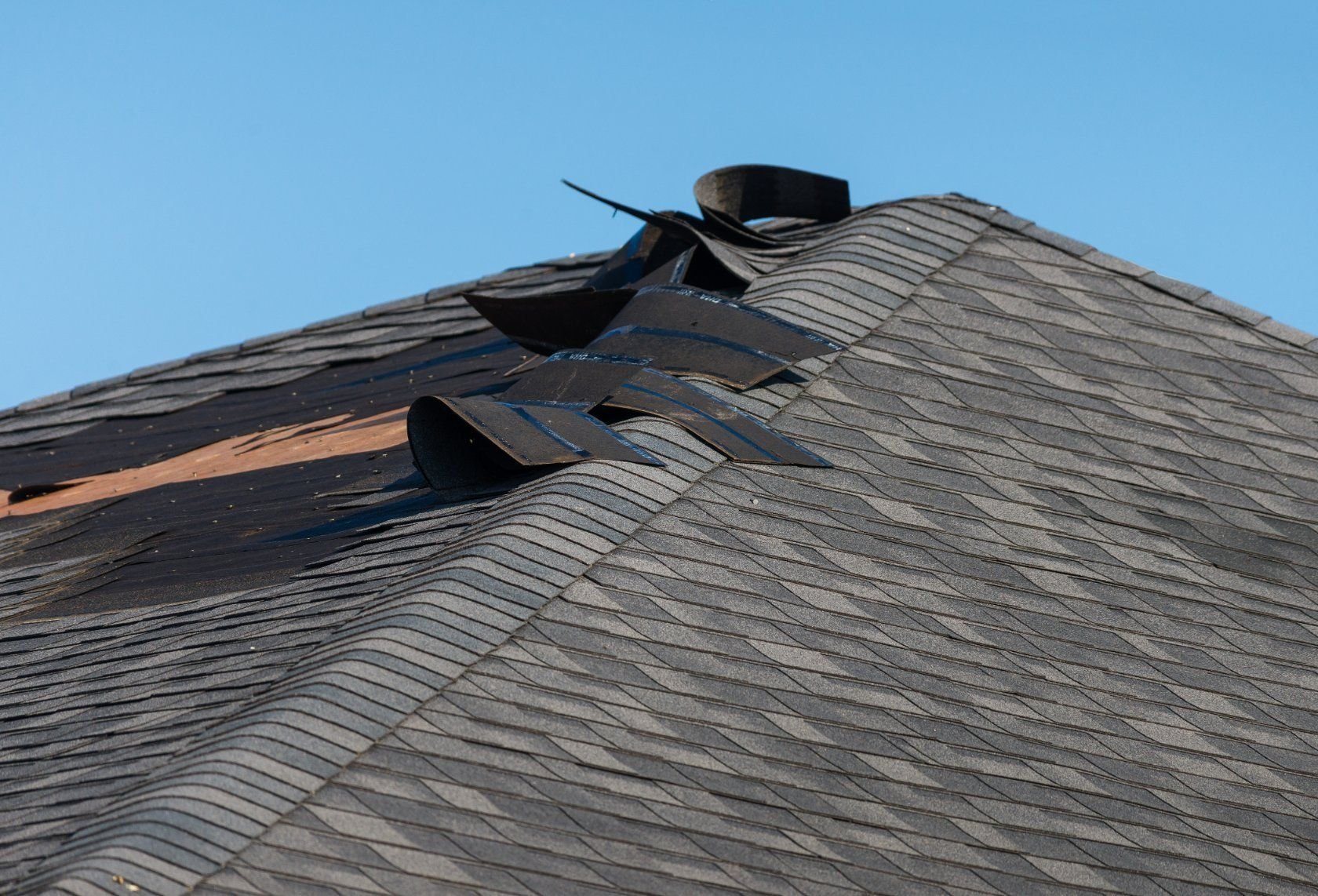 Damaged asphalt shingle roof with torn and curled material against a blue sky