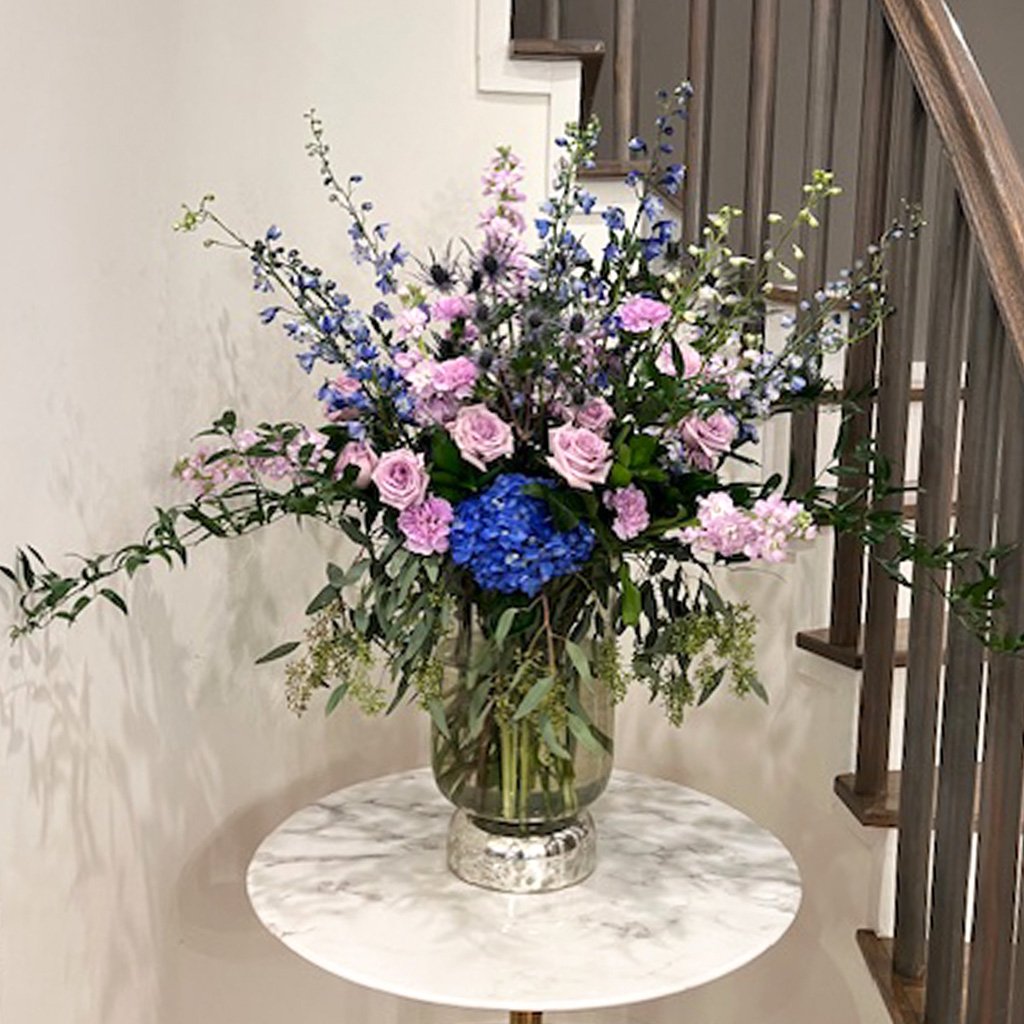 A tall vase of pink and blue flowers on a marble table near a staircase
