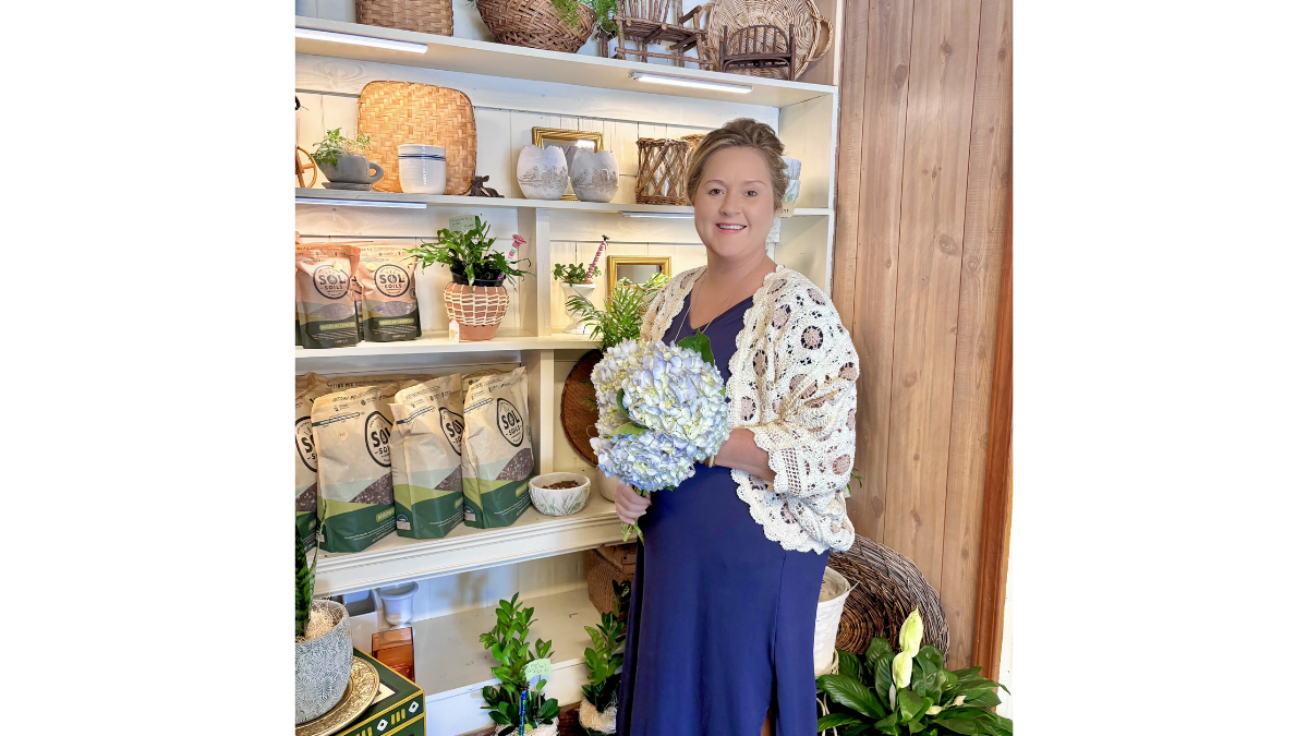 A woman in a blue dress holding a bouquet of flowers stands in a shop with shelves of plants and products.