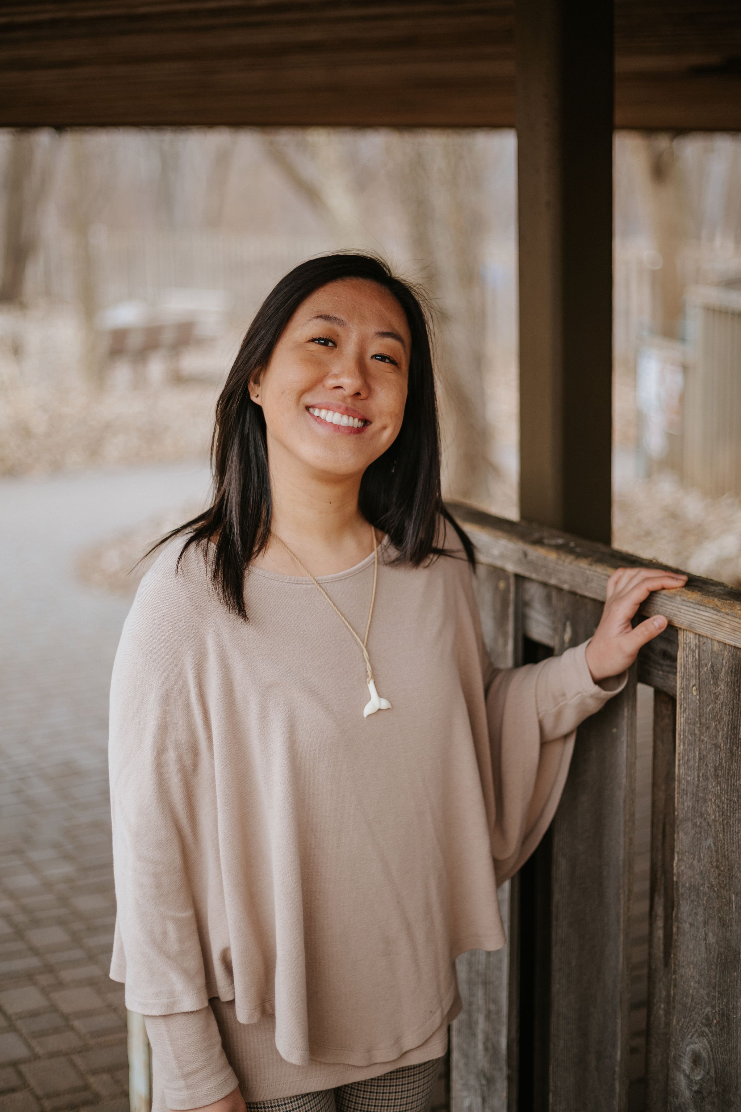 A smiling woman in a beige top stands on a wooden deck, looking out at a park.