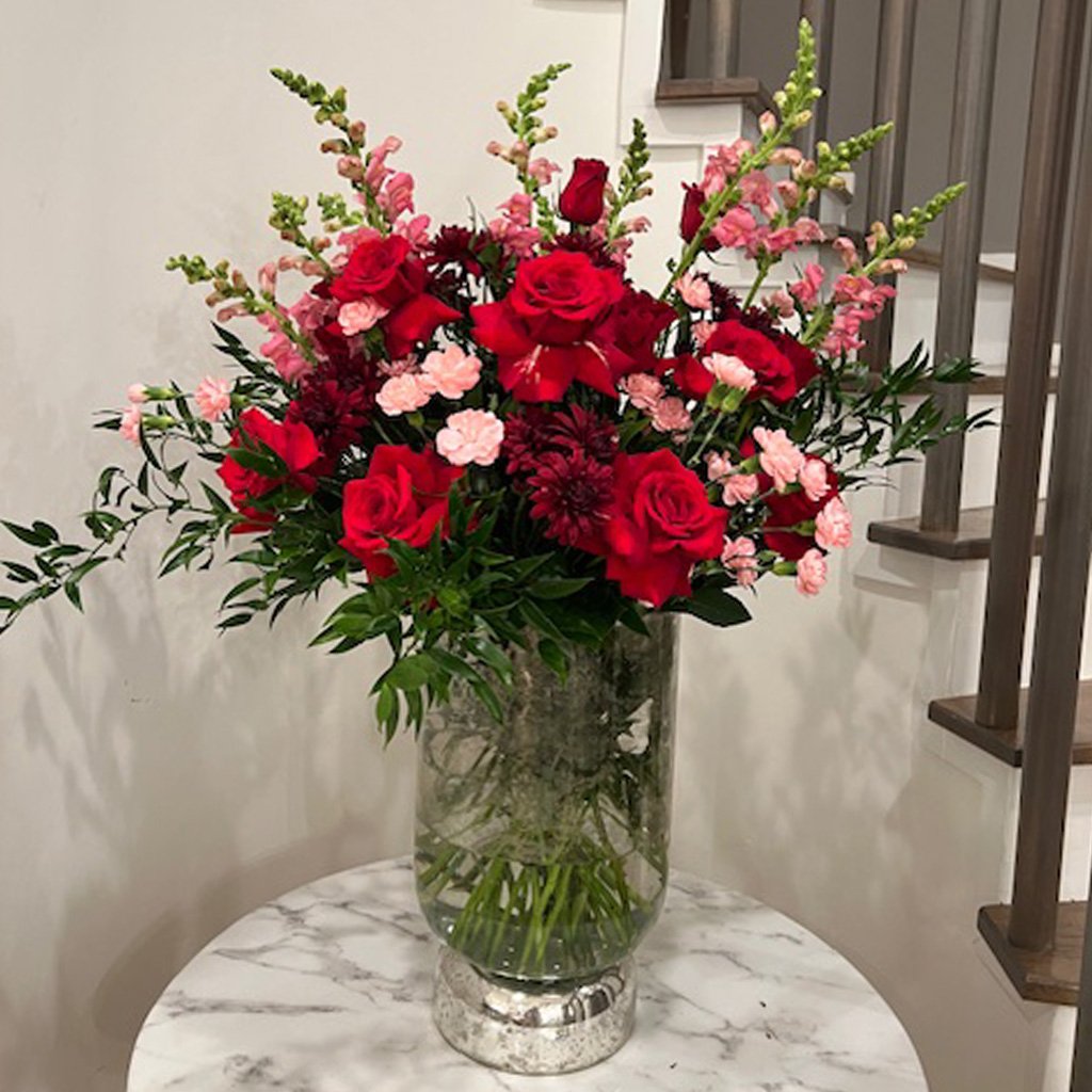 A tall vase filled with red roses, pink carnations, and green foliage on a marble table
