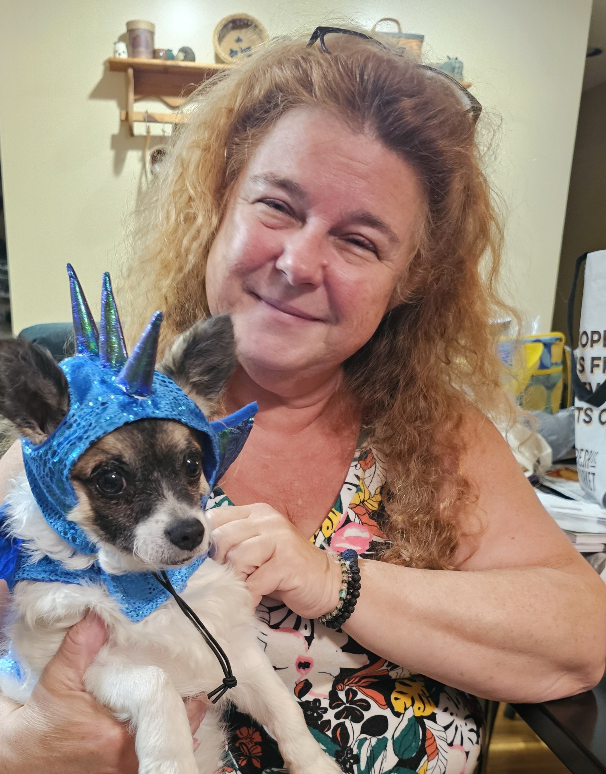 A woman with curly hair holds a small dog wearing a blue unicorn hat indoors.