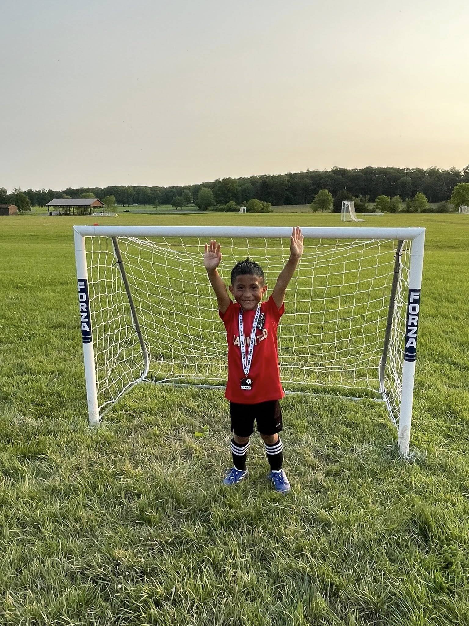 A young boy in a red shirt stands in front of a soccer goal with a medal