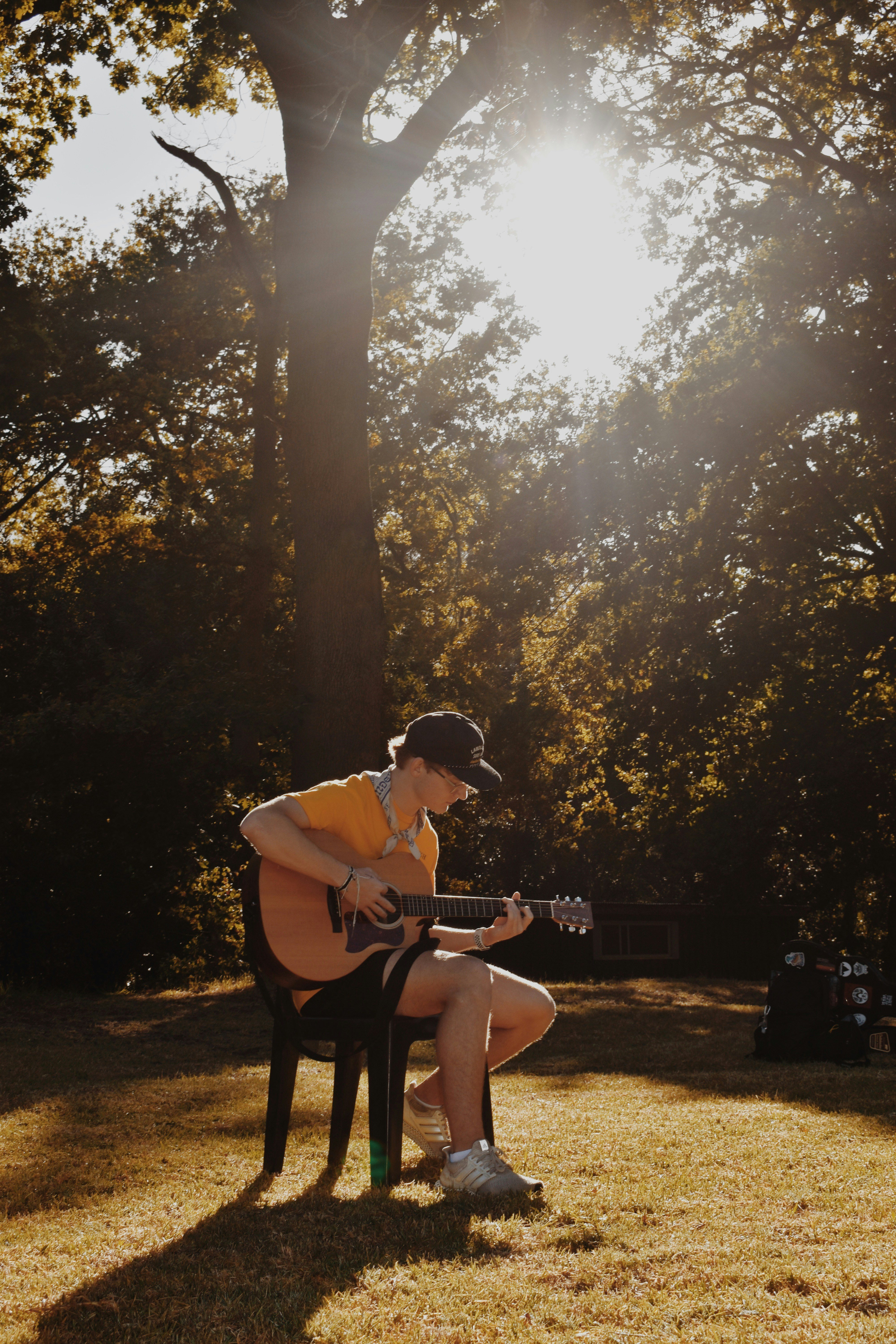 A man in a yellow shirt and black hat plays a guitar in a park under a tree.