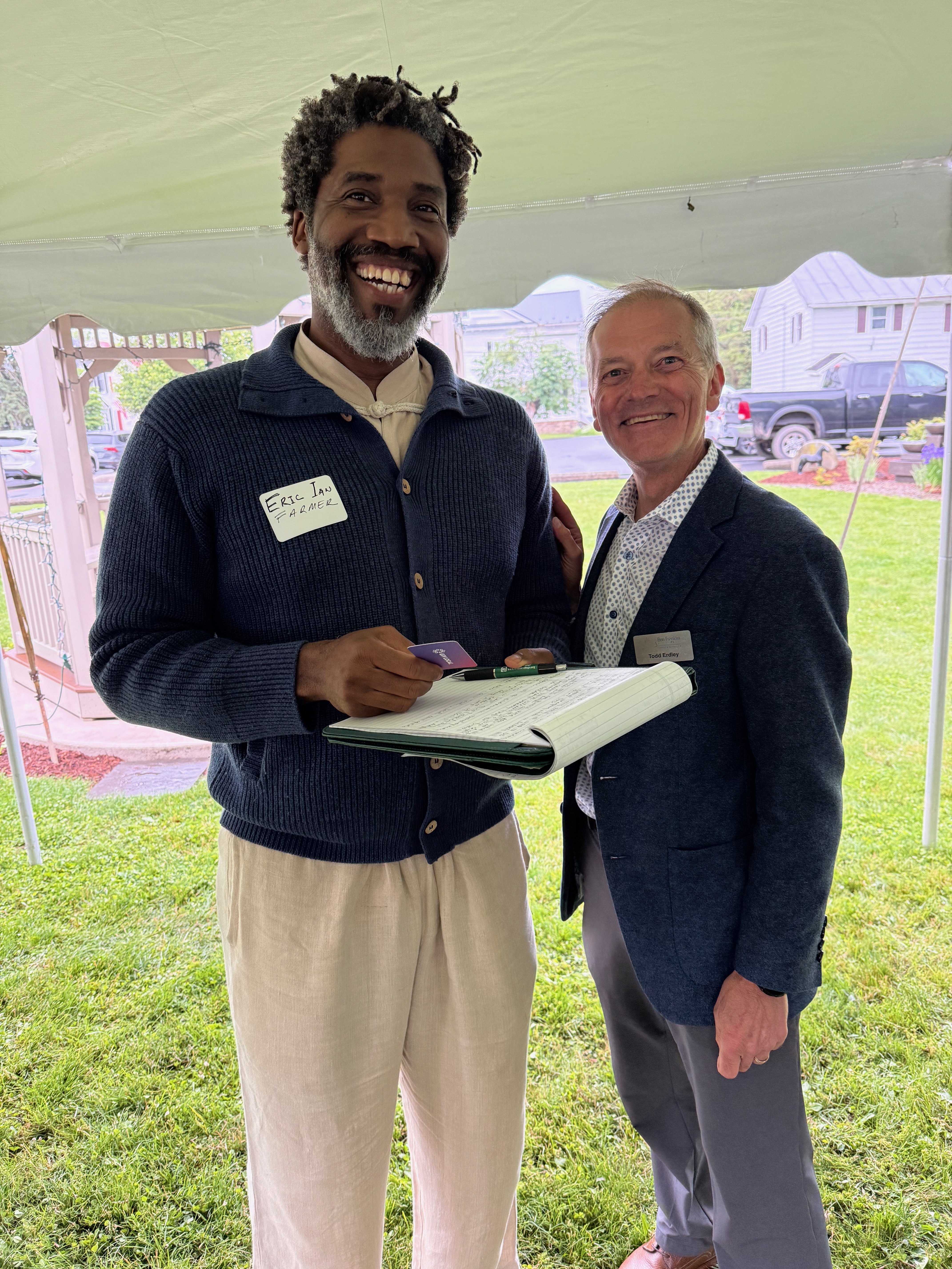 Two men smiling and standing under a tent, one holding a clipboard and pen
