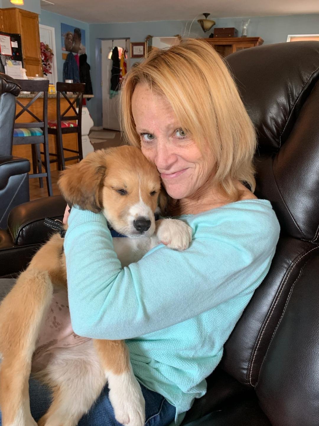 A woman with blonde hair is sitting on a black leather couch and hugging a brown and white dog.