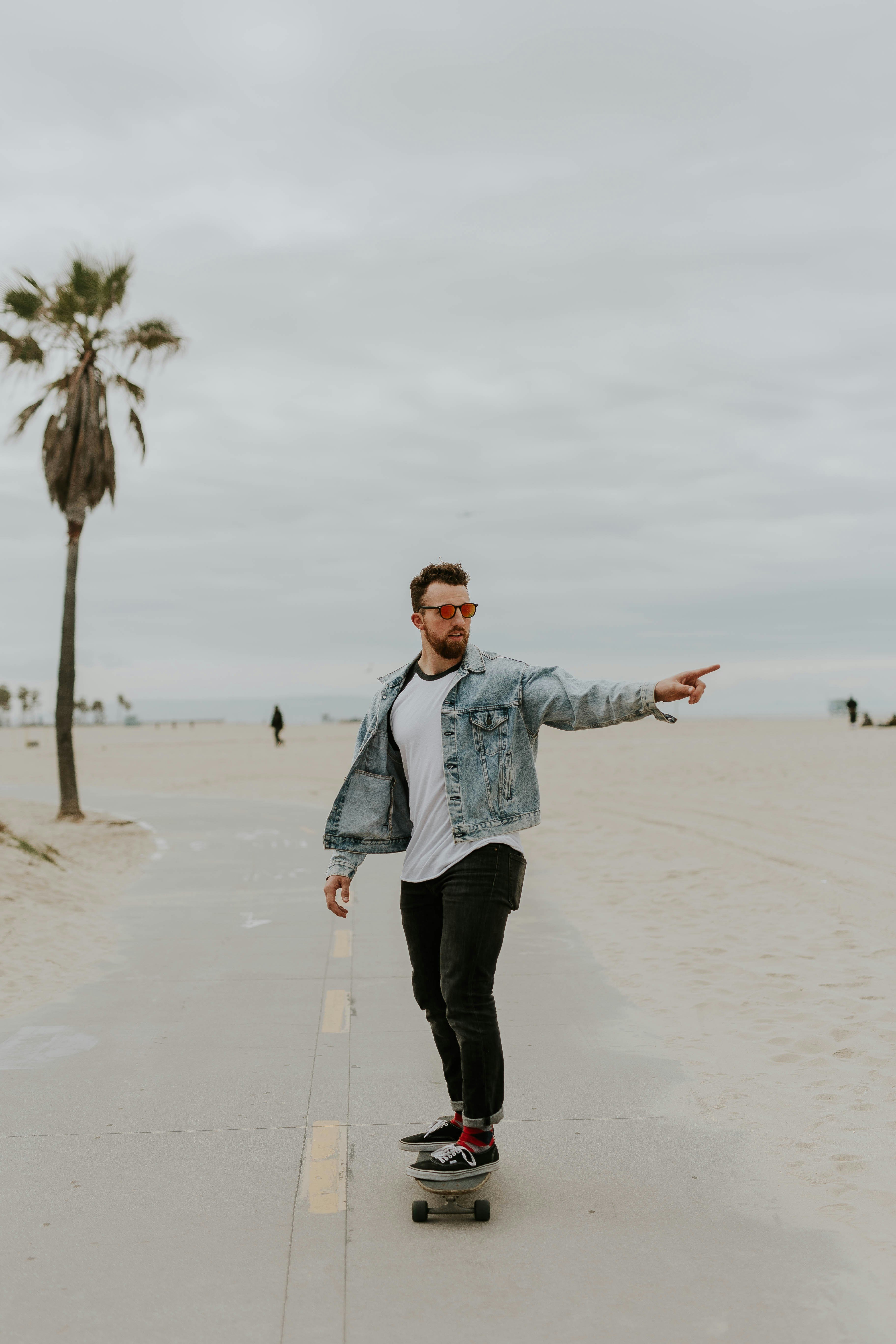 A man wearing sunglasses and a denim jacket rides a skateboard on a beach path.