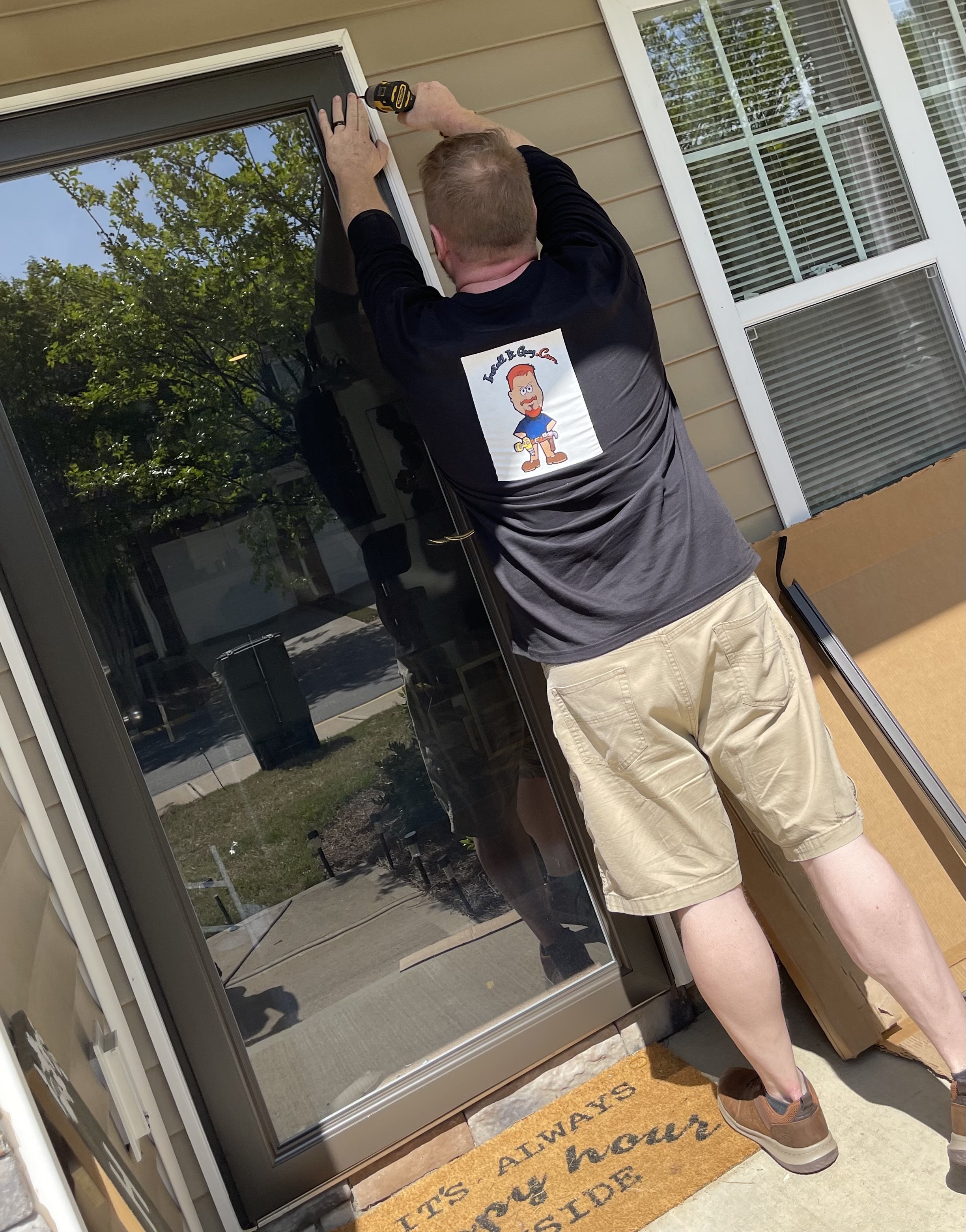 A man in a gray shirt with a cartoon character on it is fixing a glass door.
