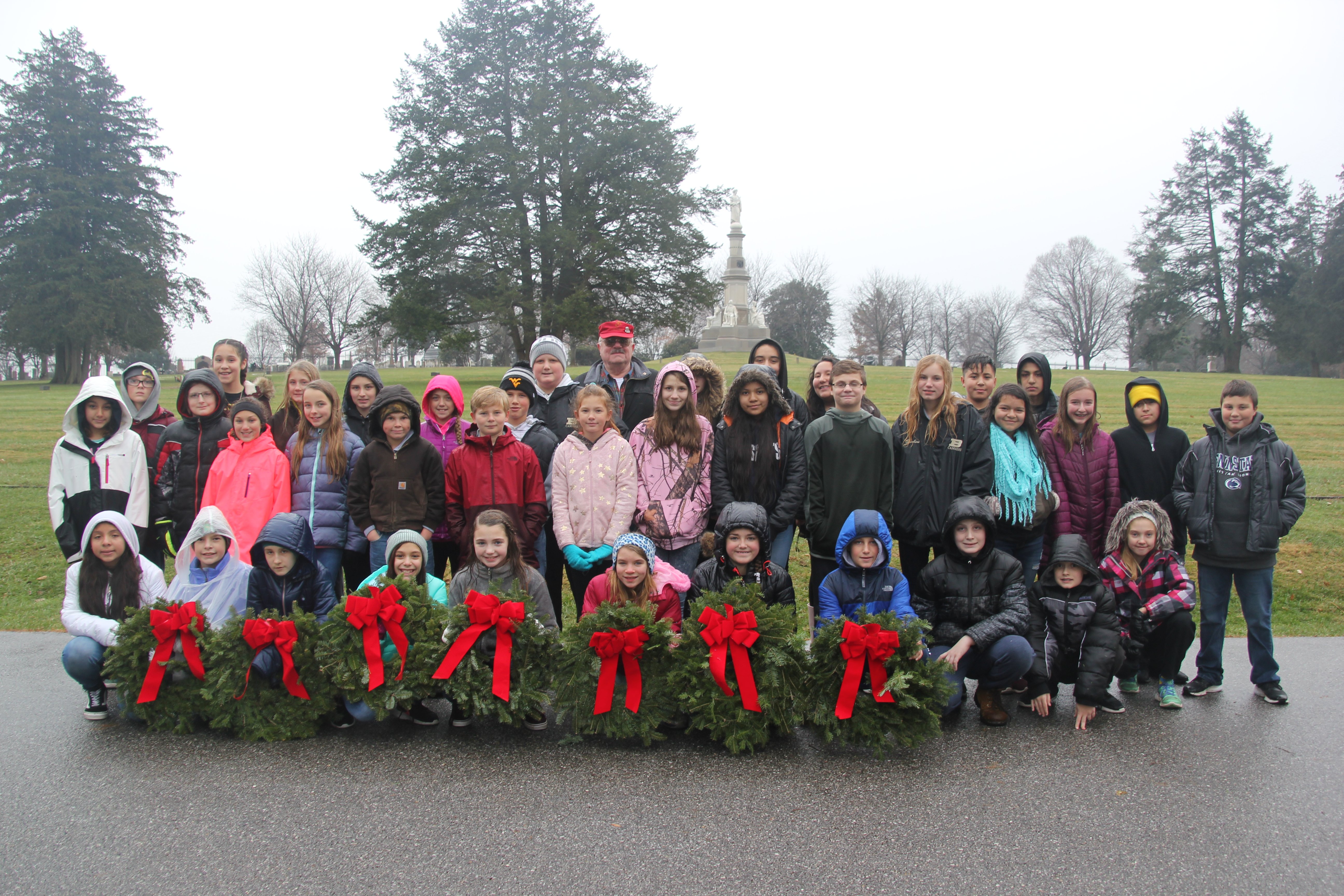 A group of students and teachers pose with wreaths in front of a memorial.