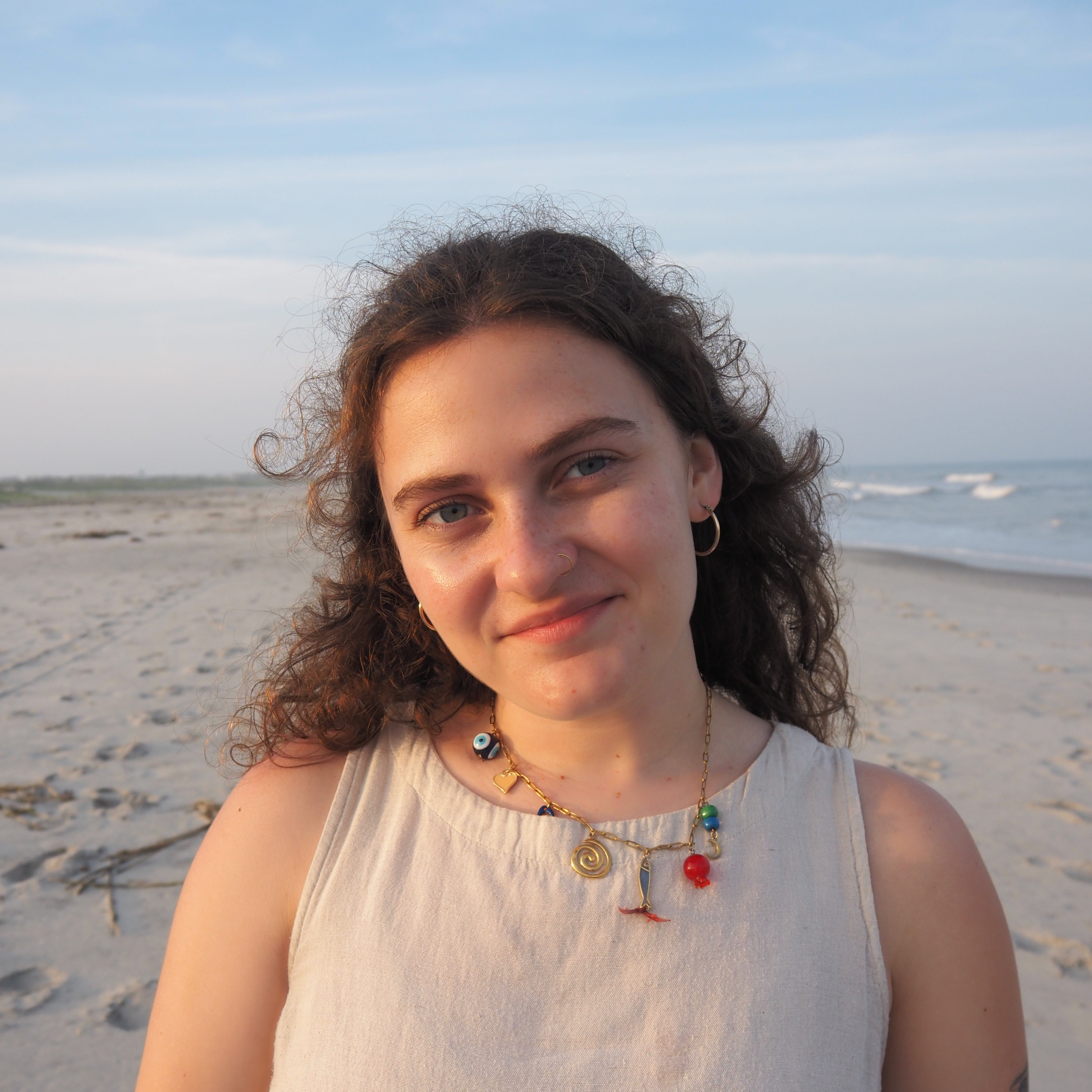 A woman with curly hair and a nose ring smiles on a beach with the ocean in the background.