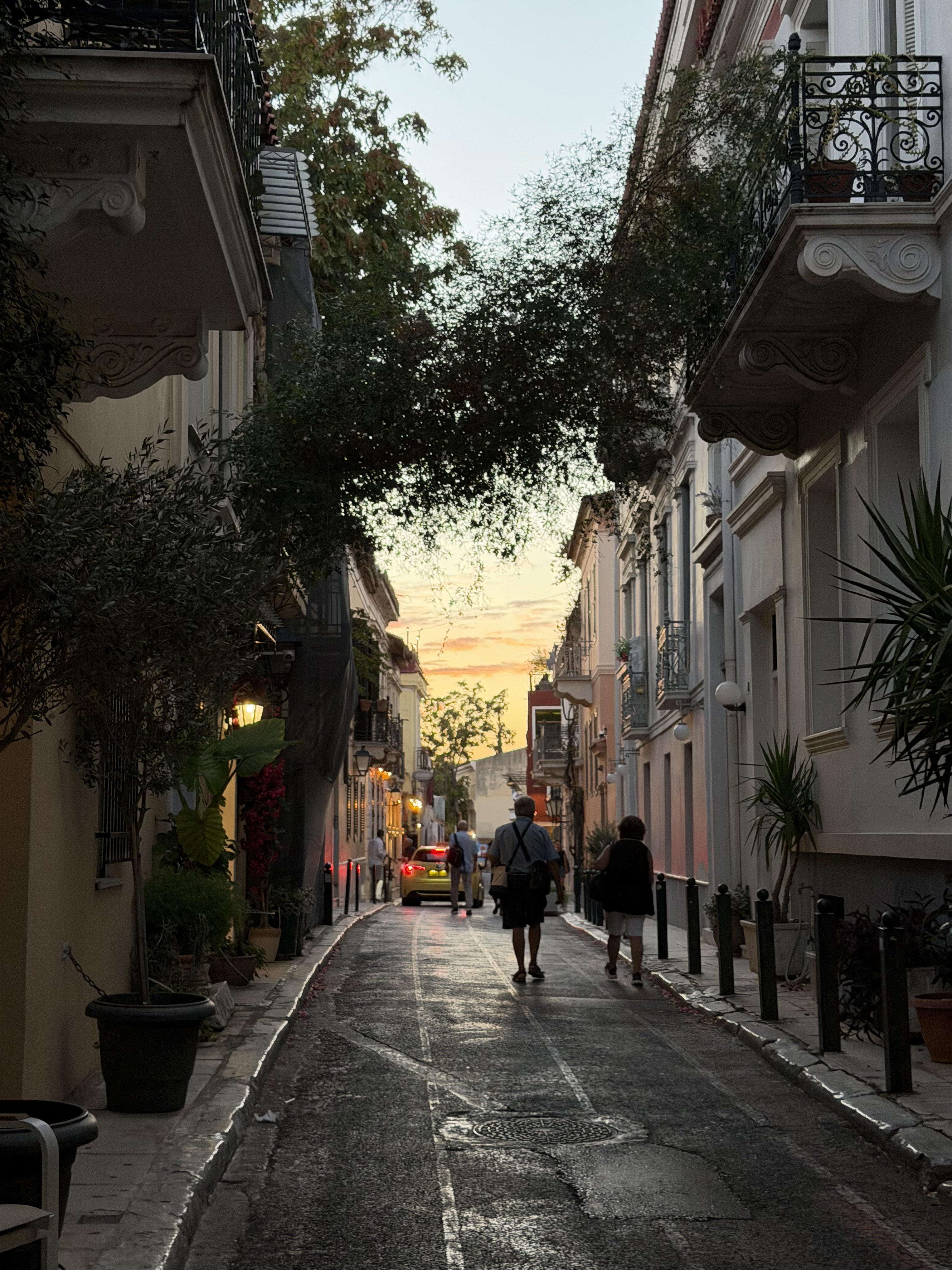 A narrow street with people walking and a car driving by at sunset