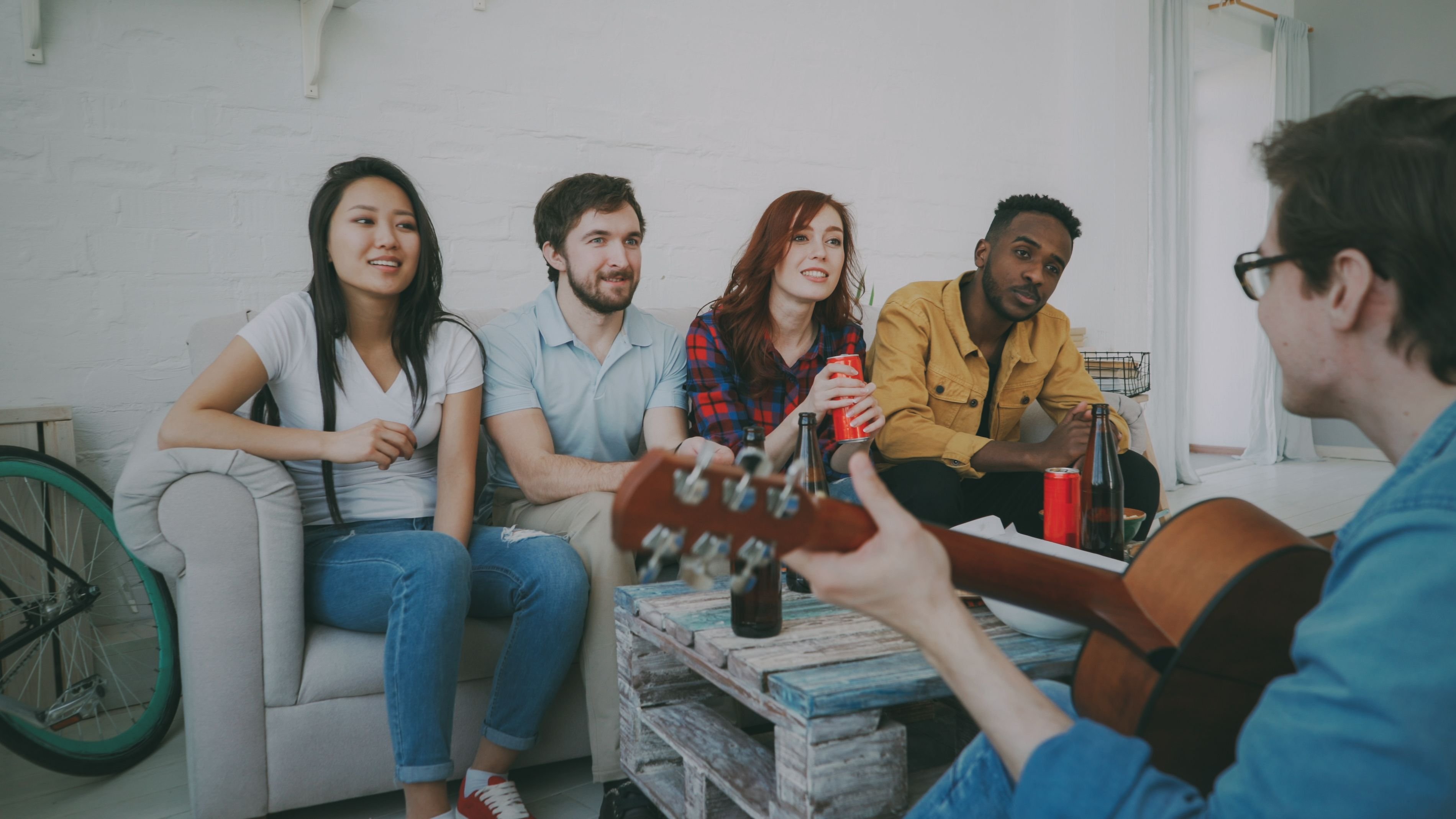 Five friends sitting on a couch, one playing guitar, smiling and chatting