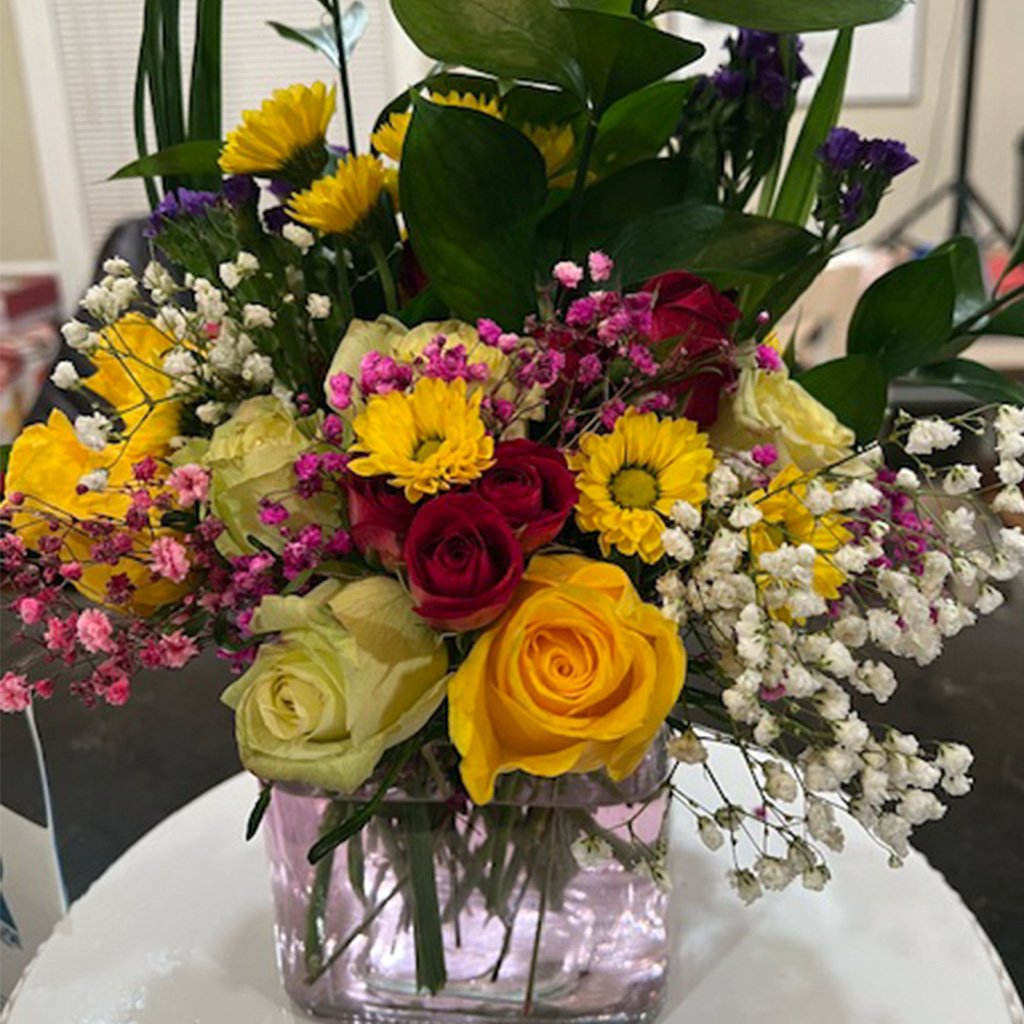 A vibrant bouquet of roses, daisies, and other flowers in a square glass vase