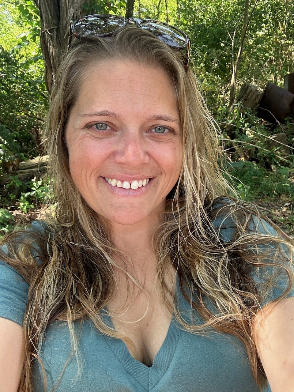 A woman with blonde hair and glasses on her head smiles in a forest.
