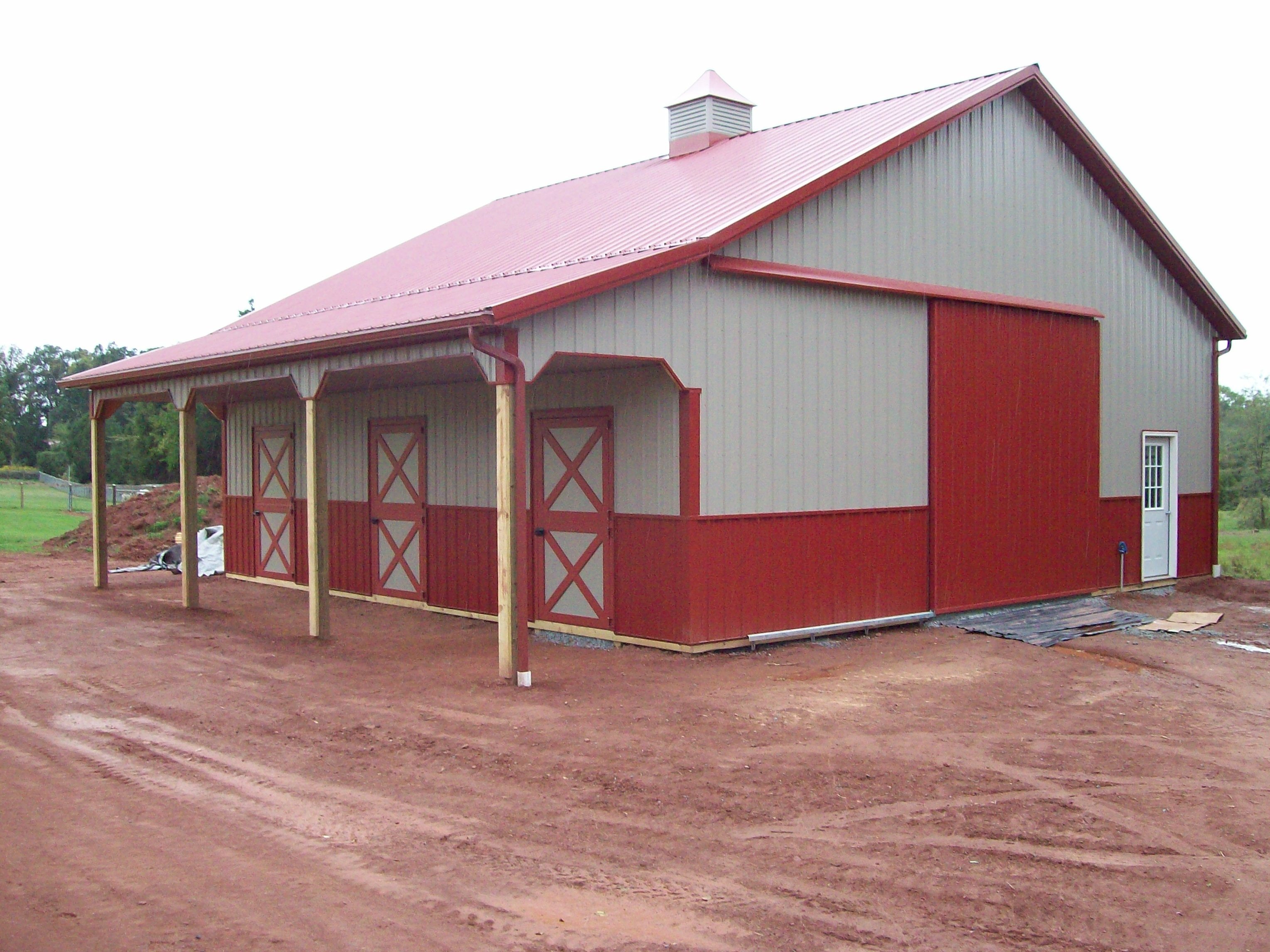 A red and gray barn with a red roof and three doors, under construction with dirt ground