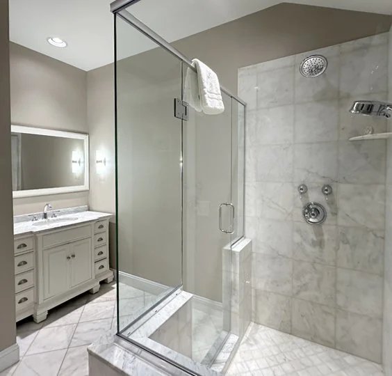 A modern bathroom with a glass shower enclosure, white marble tiles, and a white vanity with drawers.